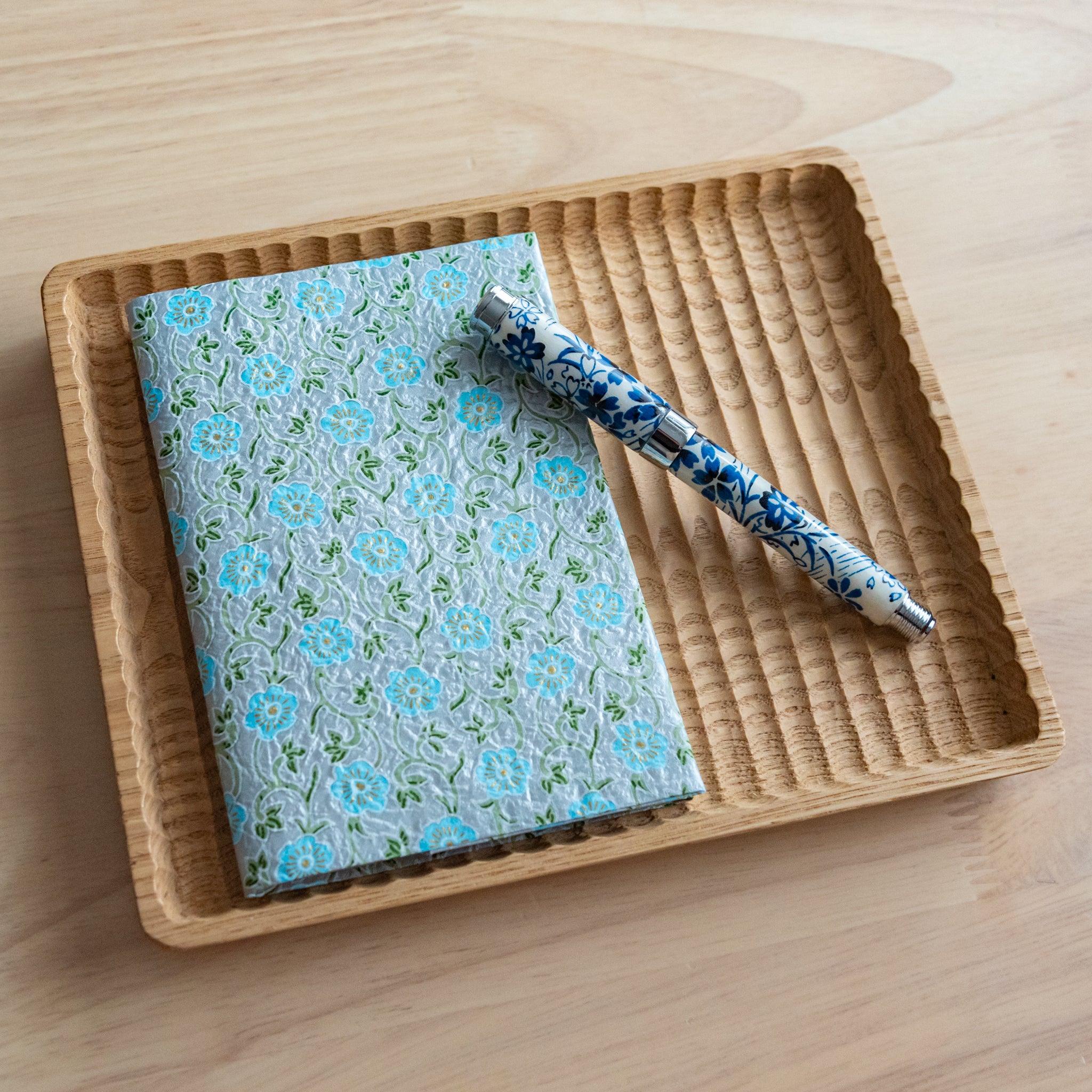 A notebook with a blue floral pattern and a matching blue and white pen, neatly placed on a wooden tray on a light wooden surface.