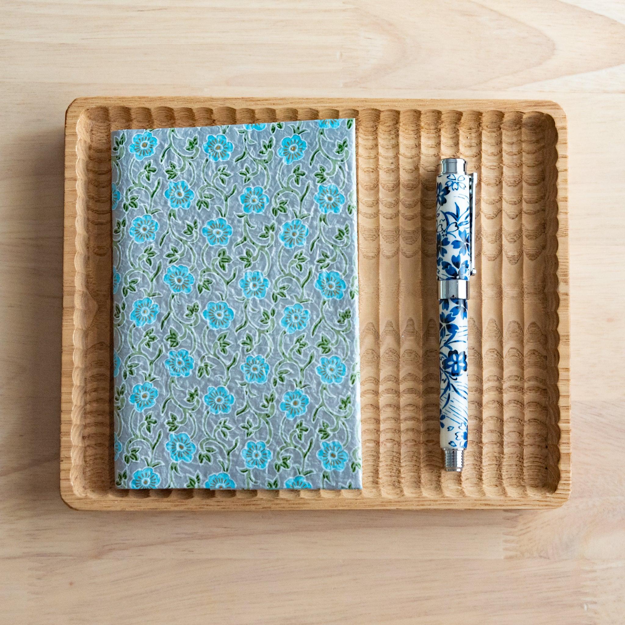 A notebook with a blue floral pattern and a matching blue and white pen, neatly arranged on a wooden tray on a light wooden surface.