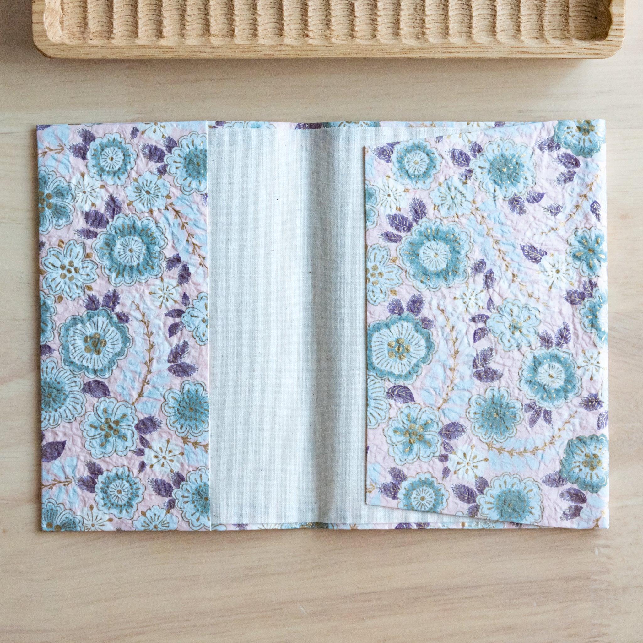 An open notebook cover with a pastel floral pattern featuring blue and purple flowers, revealing its inner white lining on a wooden surface.