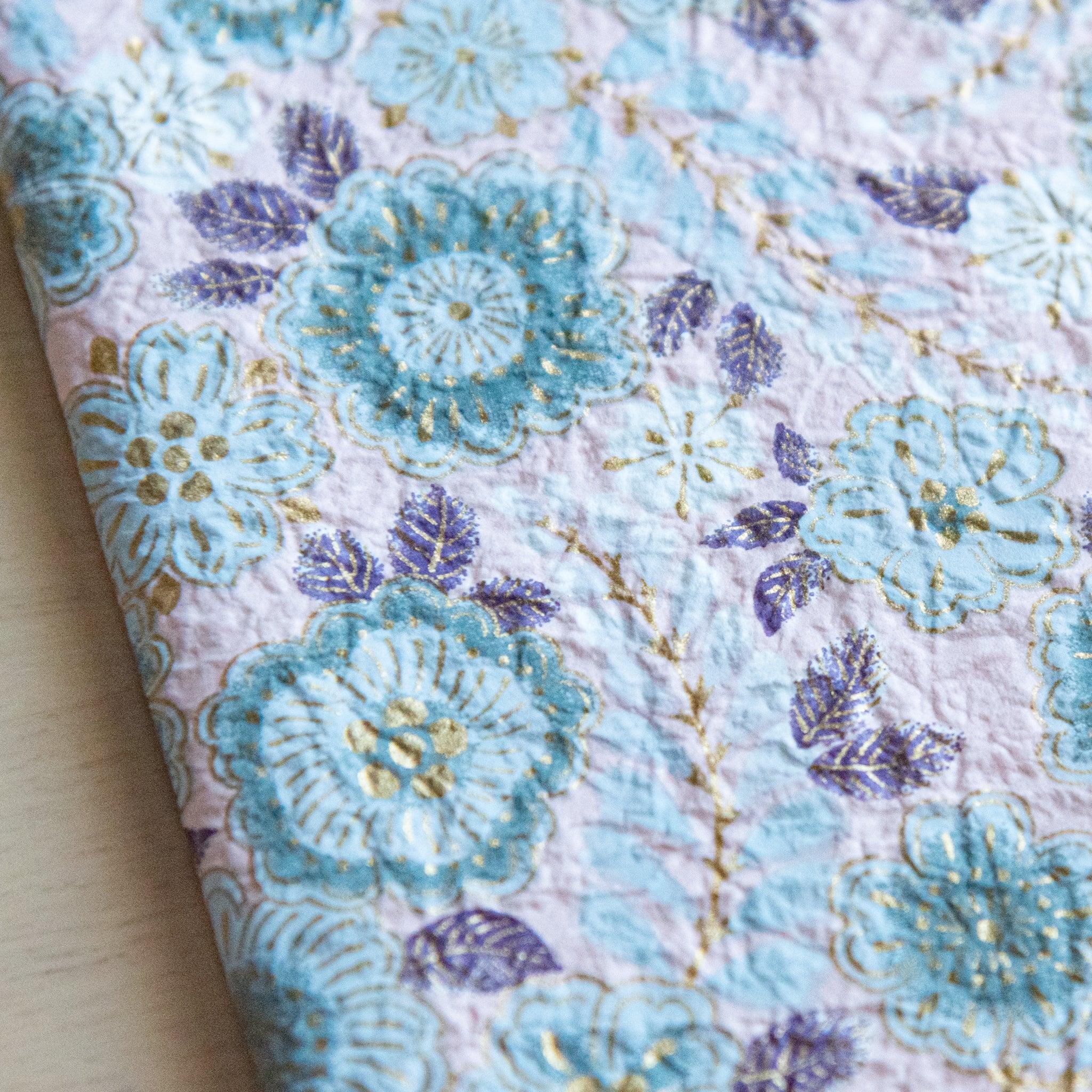 Close-up of a floral-patterned notebook cover with textured blue and gold details on a pastel pink background.