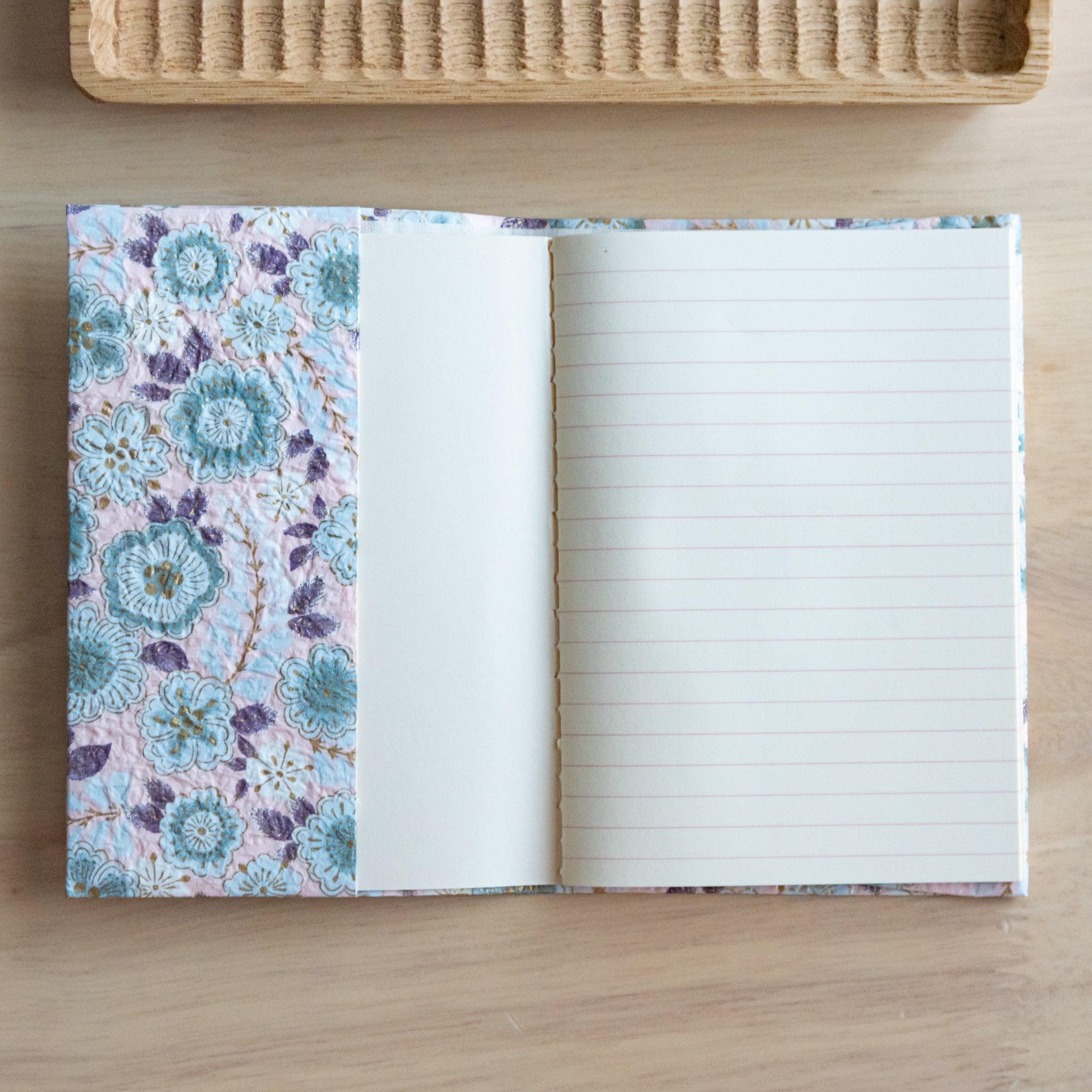 Floral-patterned notebook with an open page showing lined paper, placed on a light wooden surface with a rattan tray in the background.