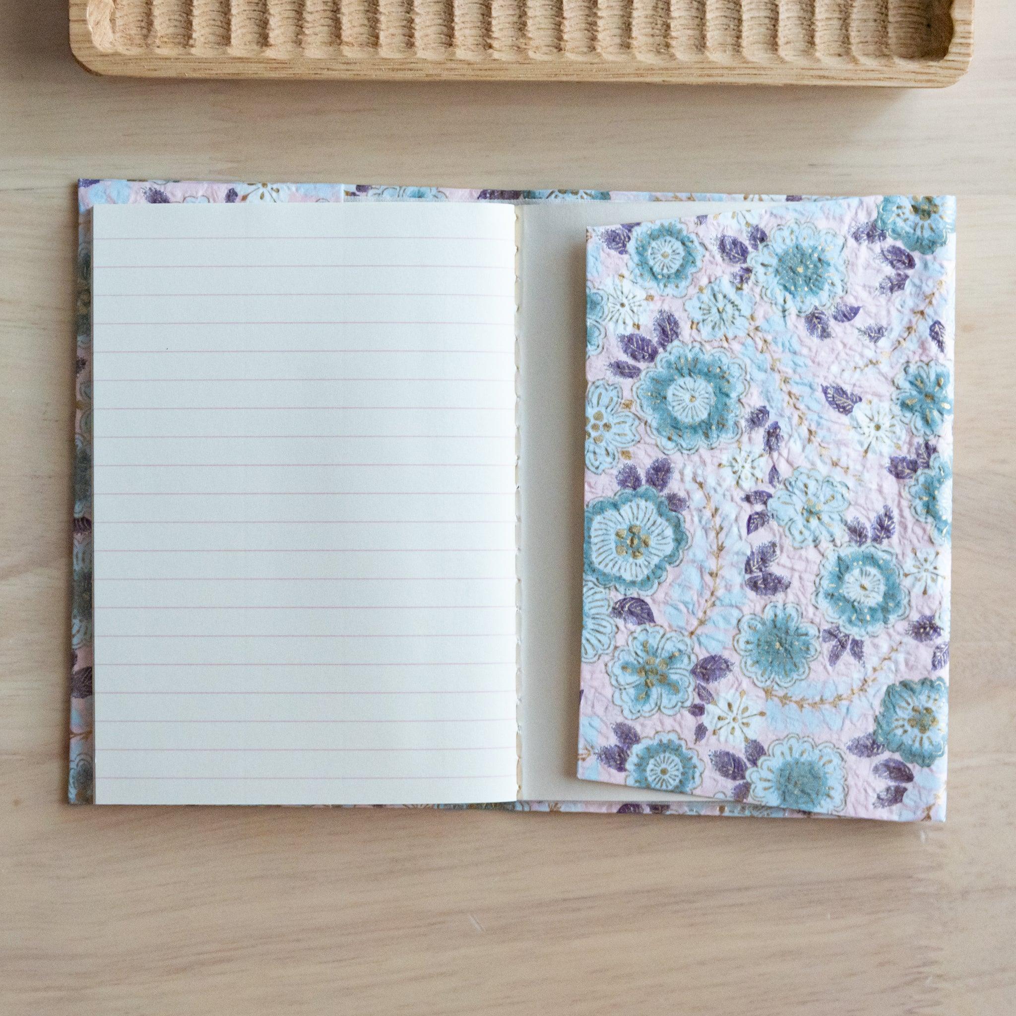 Open notebook with floral-patterned cover and lined pages, placed on a light wooden surface near a rattan tray.