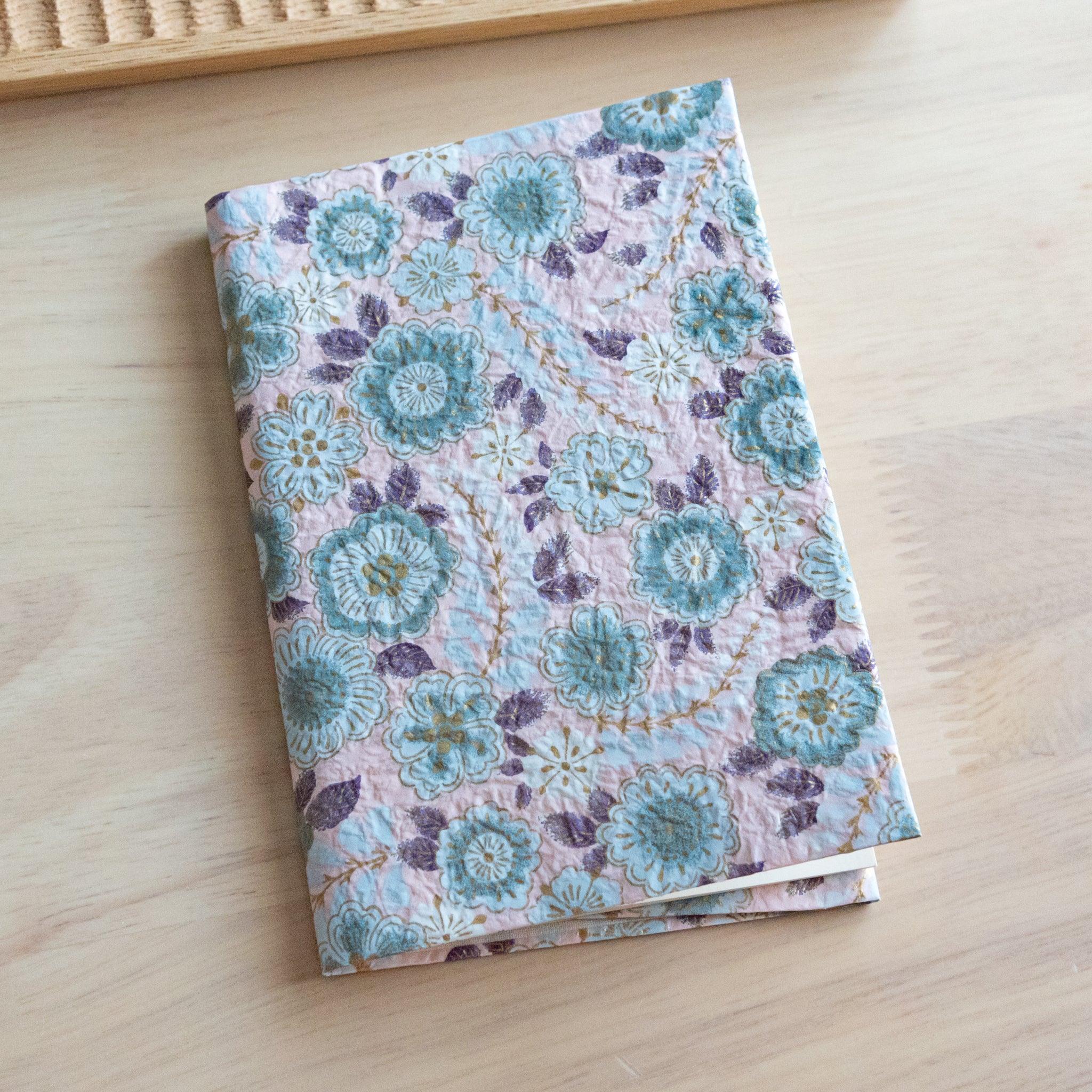 A notebook with a pastel floral pattern featuring blue and purple flowers, placed on a light wooden surface.