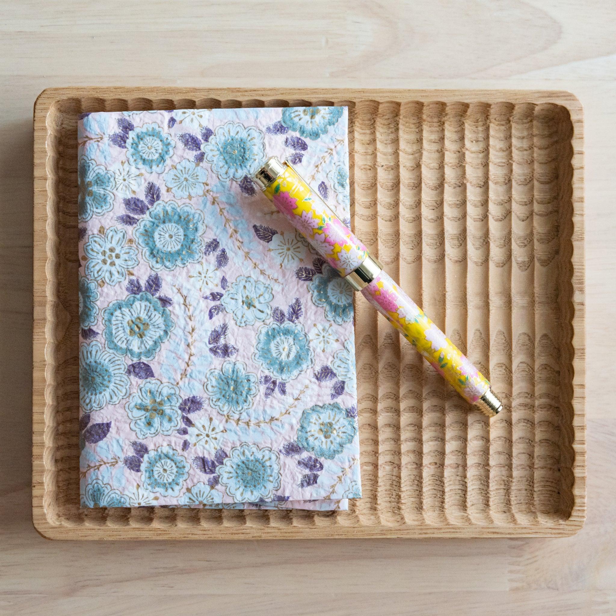 Floral-patterned notebook and a matching colorful pen placed in a rattan tray on a light wooden table.