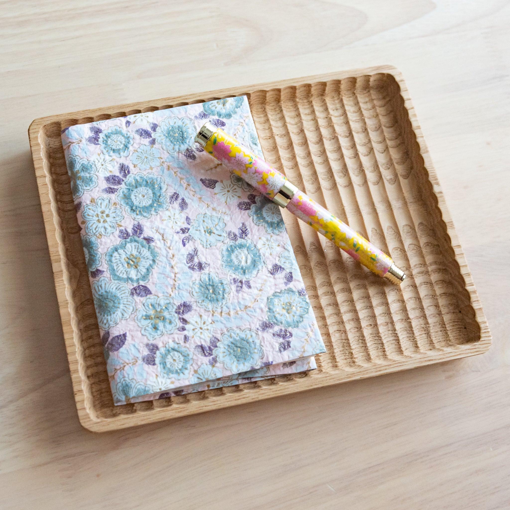 A notebook with a pastel floral pattern and a matching yellow and pink pen, neatly arranged on a wooden tray on a light wooden surface.