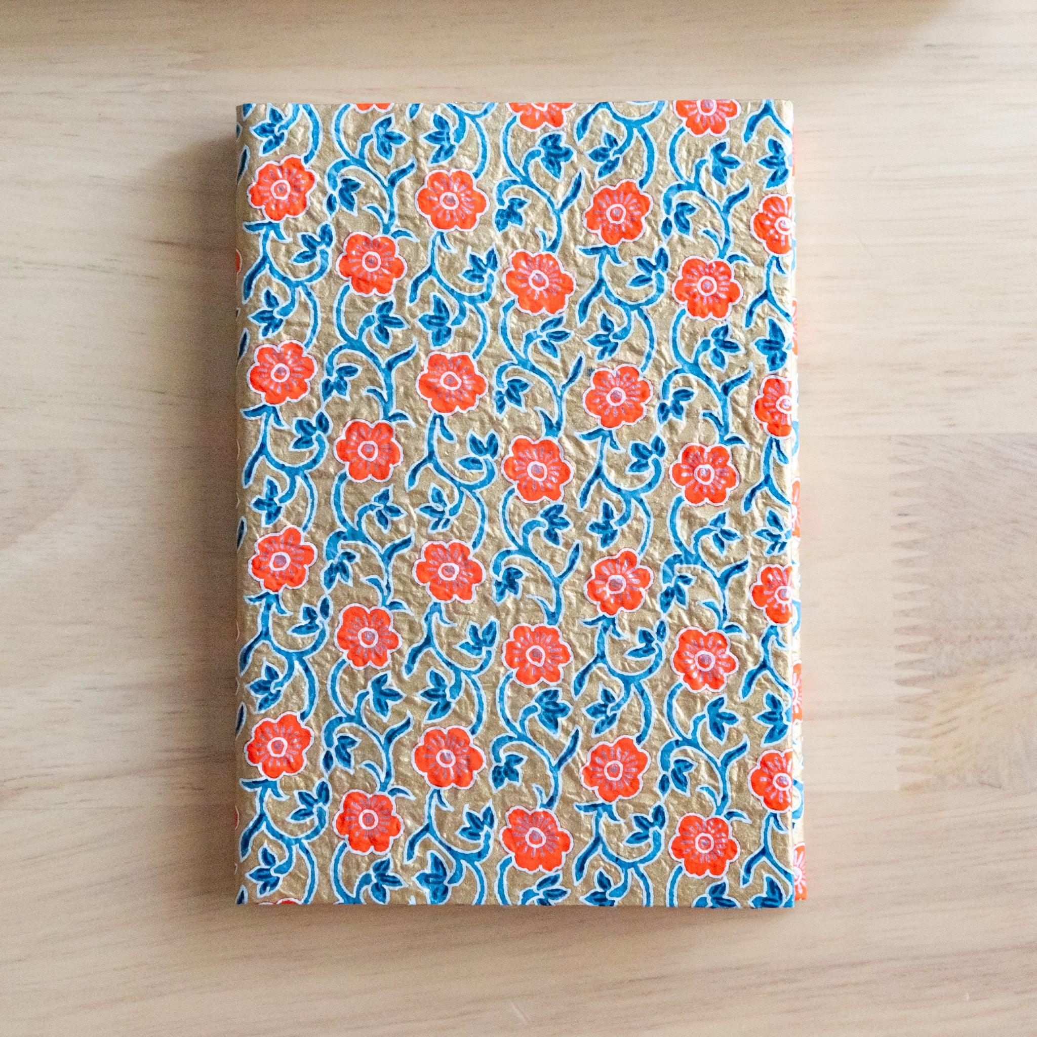 Notebook with a textured cover featuring orange flowers and blue vines on a beige background, placed on a light wooden surface.