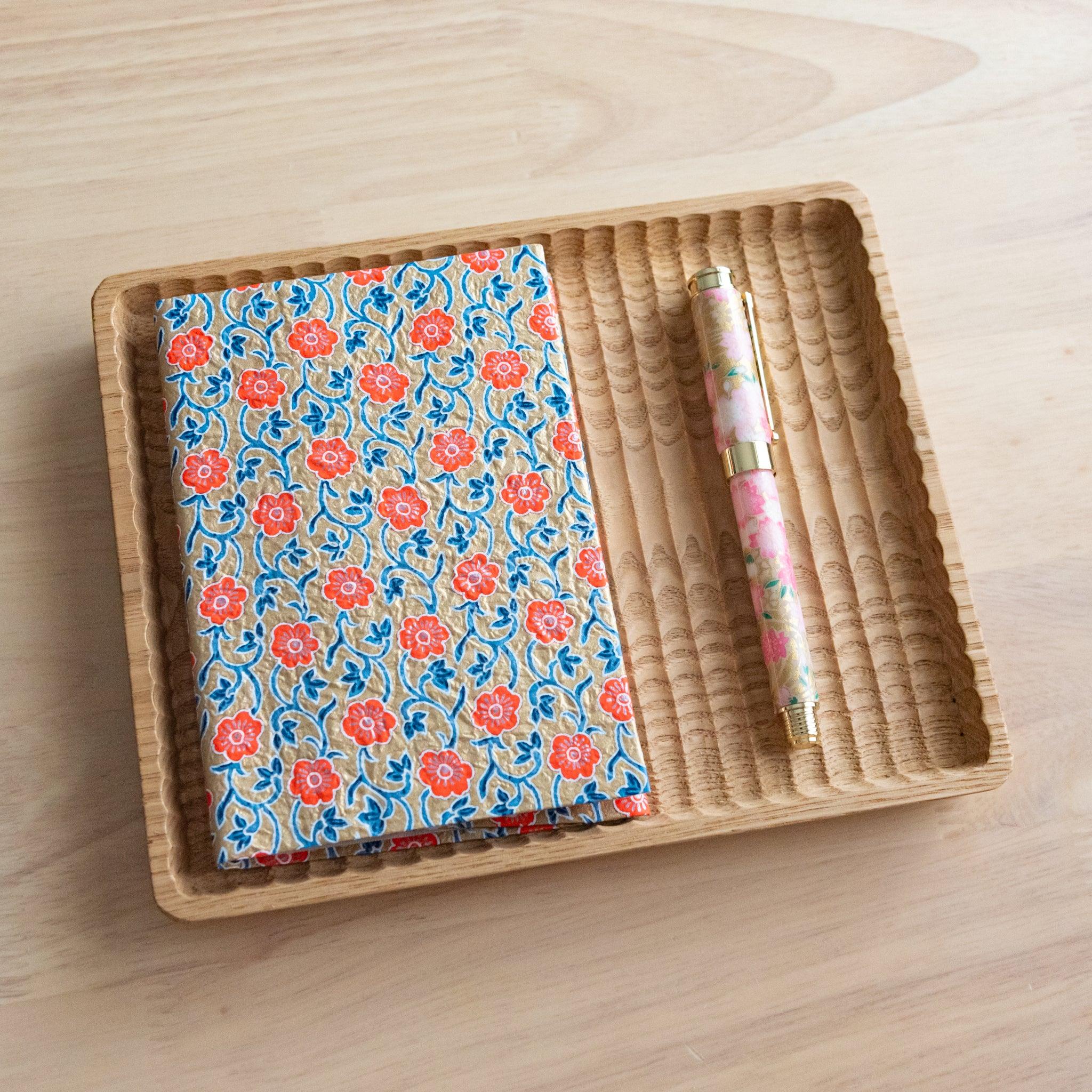 Notebook with orange floral-patterned cover and a colorful pen placed in a rattan tray on a wooden table.