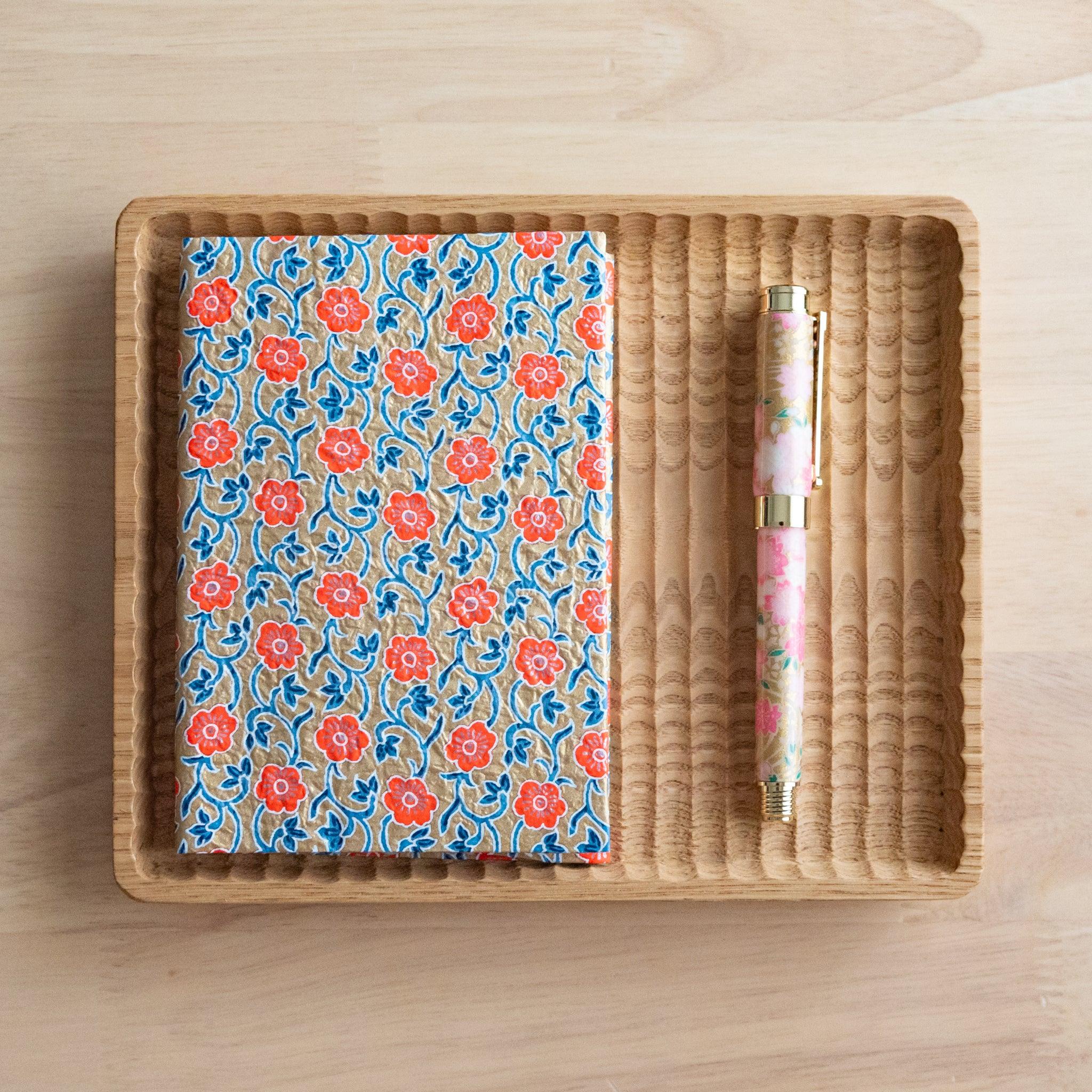 Notebook with orange floral-patterned cover and a colorful pen placed in a rattan tray on a light wooden surface.