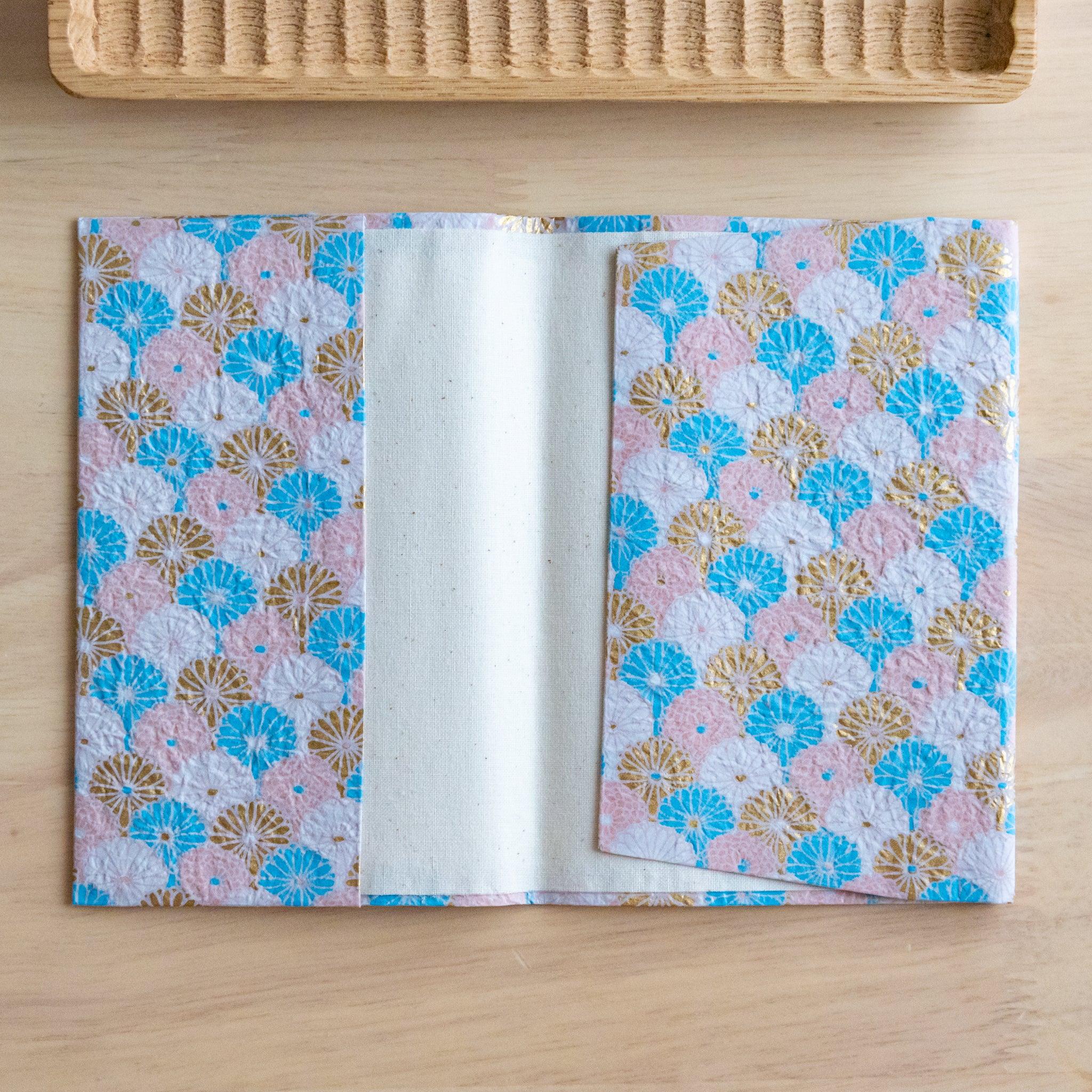 Open notebook with a pastel pink fan-patterned fabric cover showing its inner lining, placed on a wooden surface.