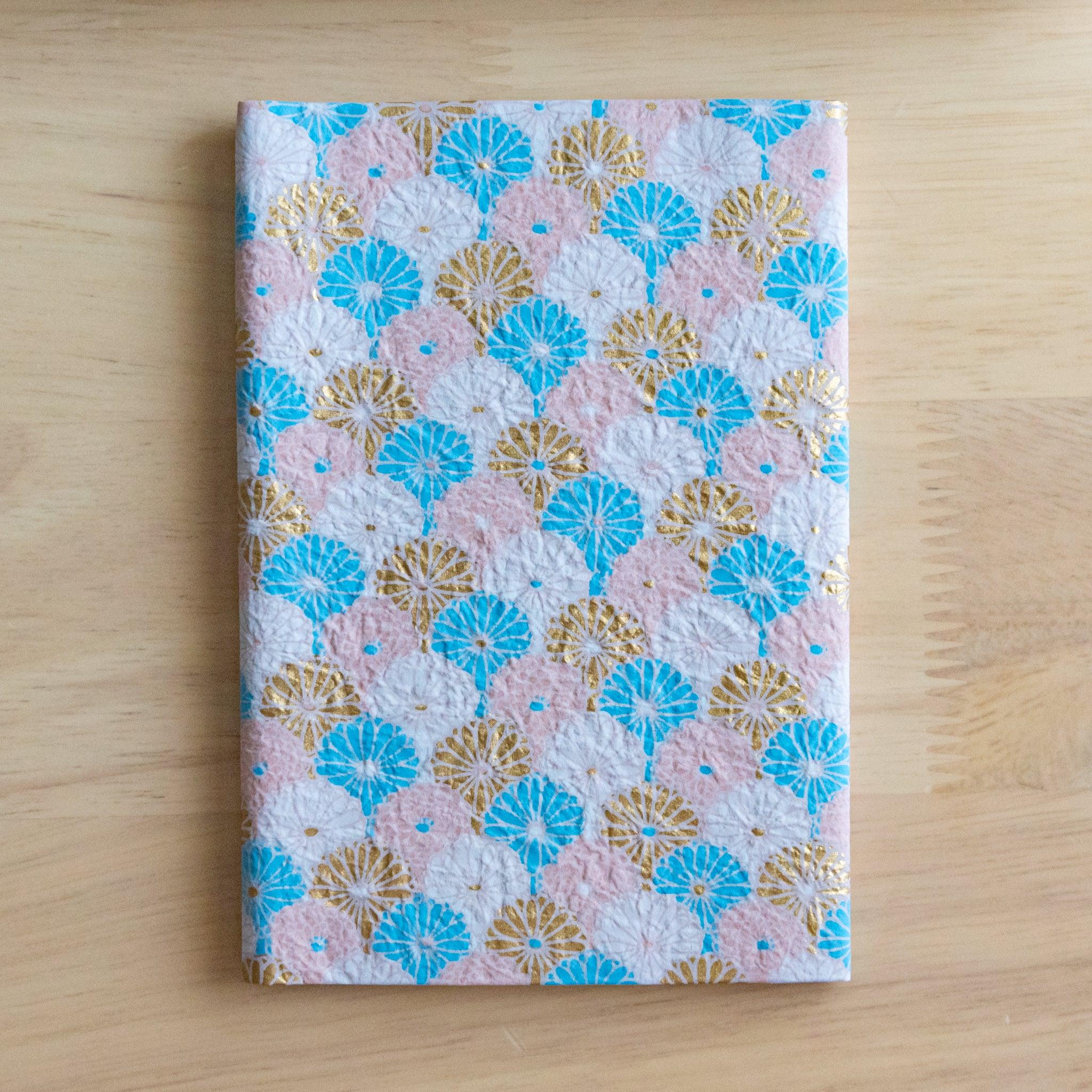 Notebook with a textured cover featuring blue and gold fan-shaped patterns on a pastel pink background, placed on a wooden surface.