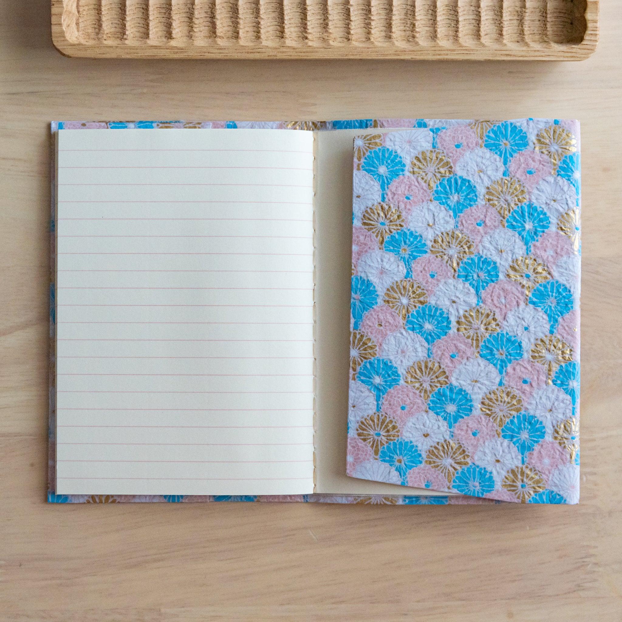 Open notebook with pastel pink and blue patterned fabric cover and lined pages, placed on a wooden surface.