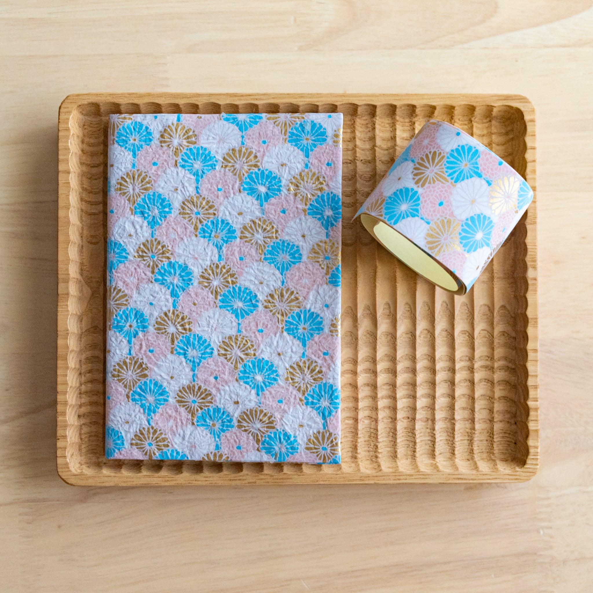 Notebook with pastel pink and blue fan-patterned cover placed in a rattan tray alongside matching decorative tape.