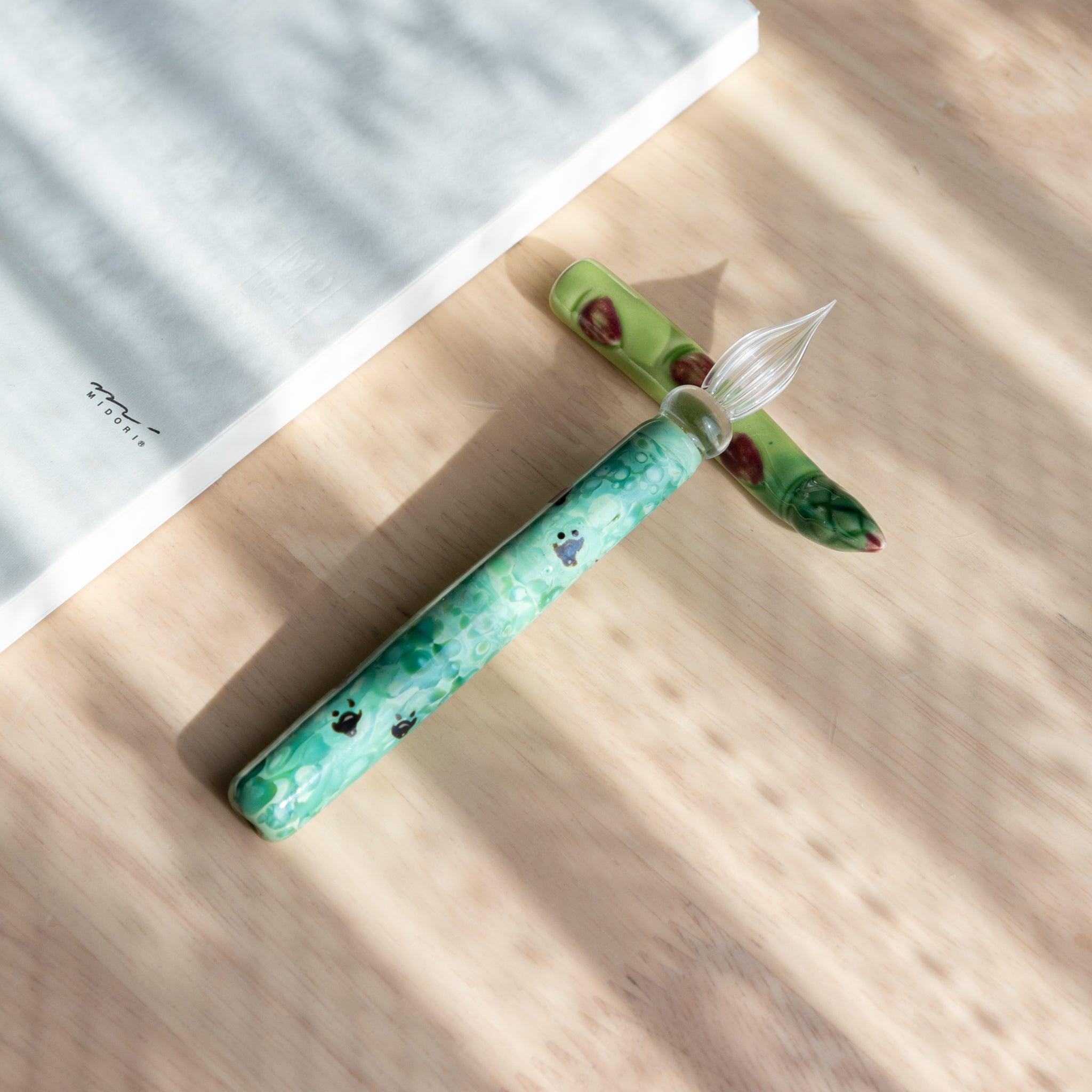 A handmade green ceramic pen rest shaped as an asparagus, holding a green glass dip pen. Part of traditional Japanese stationery items.