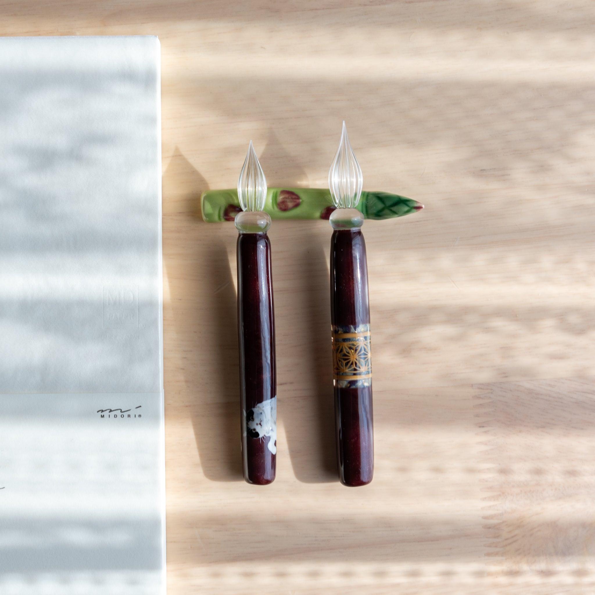 A handmade green ceramic pen rest shaped as an asparagus, holding two burgundy glass dip pens. Part of traditional Japanese stationery items.