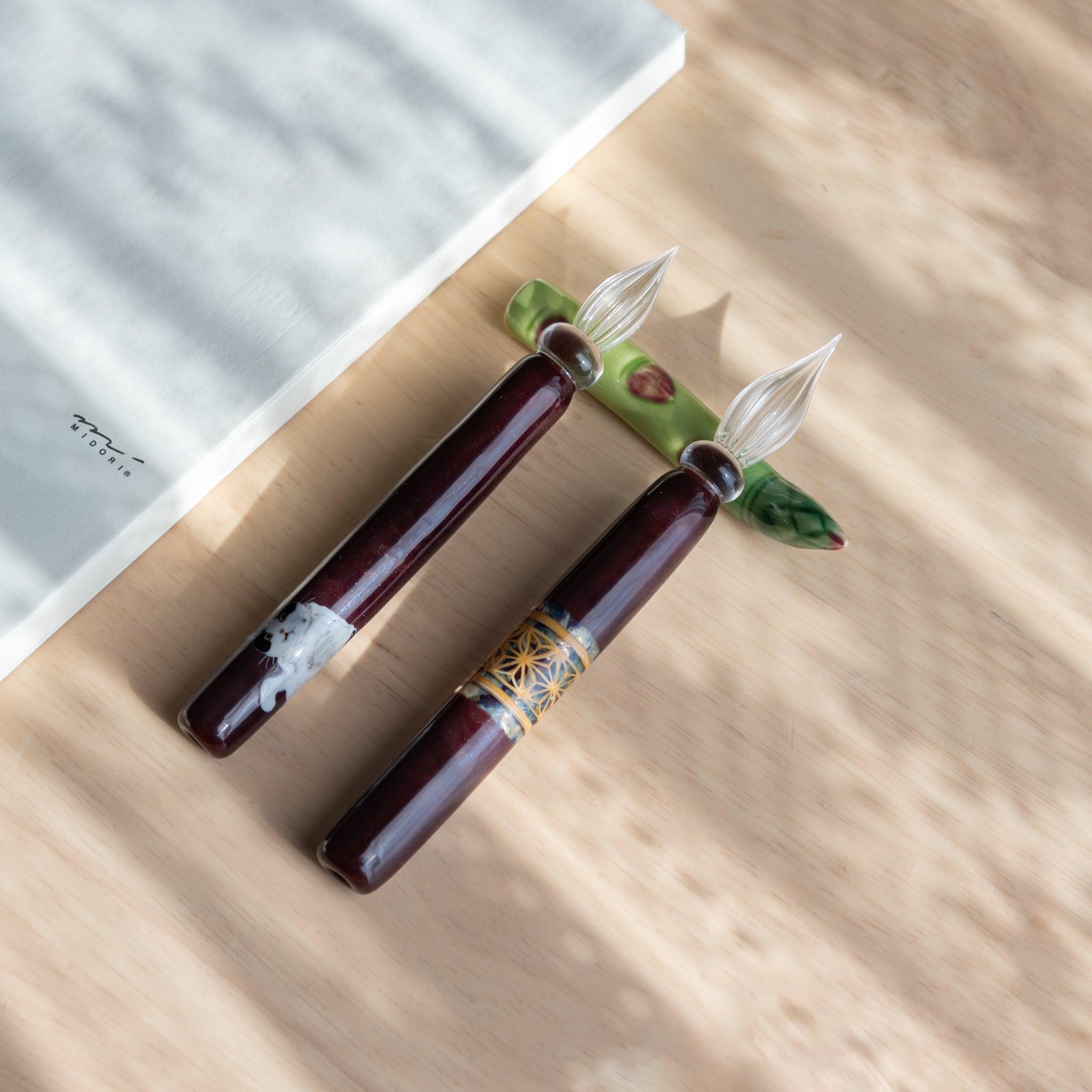 A handmade green ceramic pen rest shaped as an asparagus, holding two burgundy glass dip pens. Part of traditional Japanese stationery items.