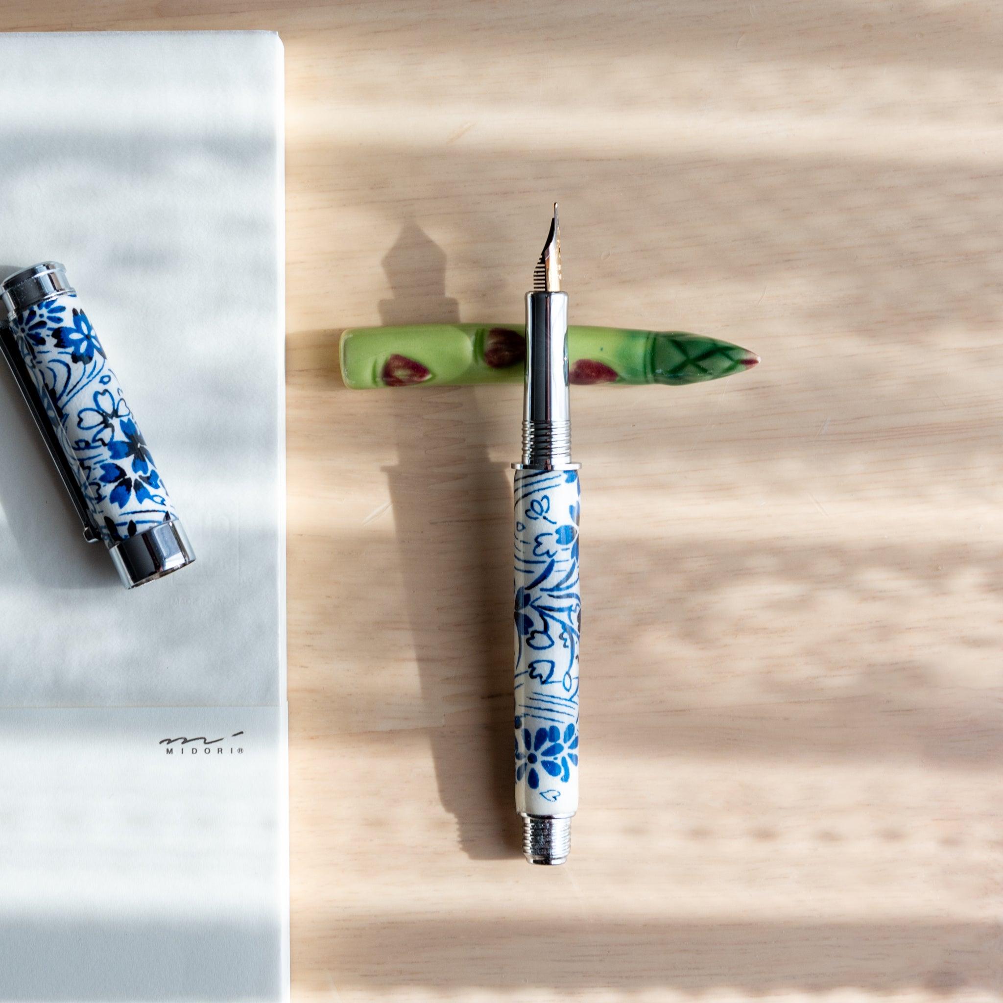 A handmade green ceramic pen rest shaped as an asparagus, holding a blue and white porcelain fountain pen. Part of Japanese stationery items.