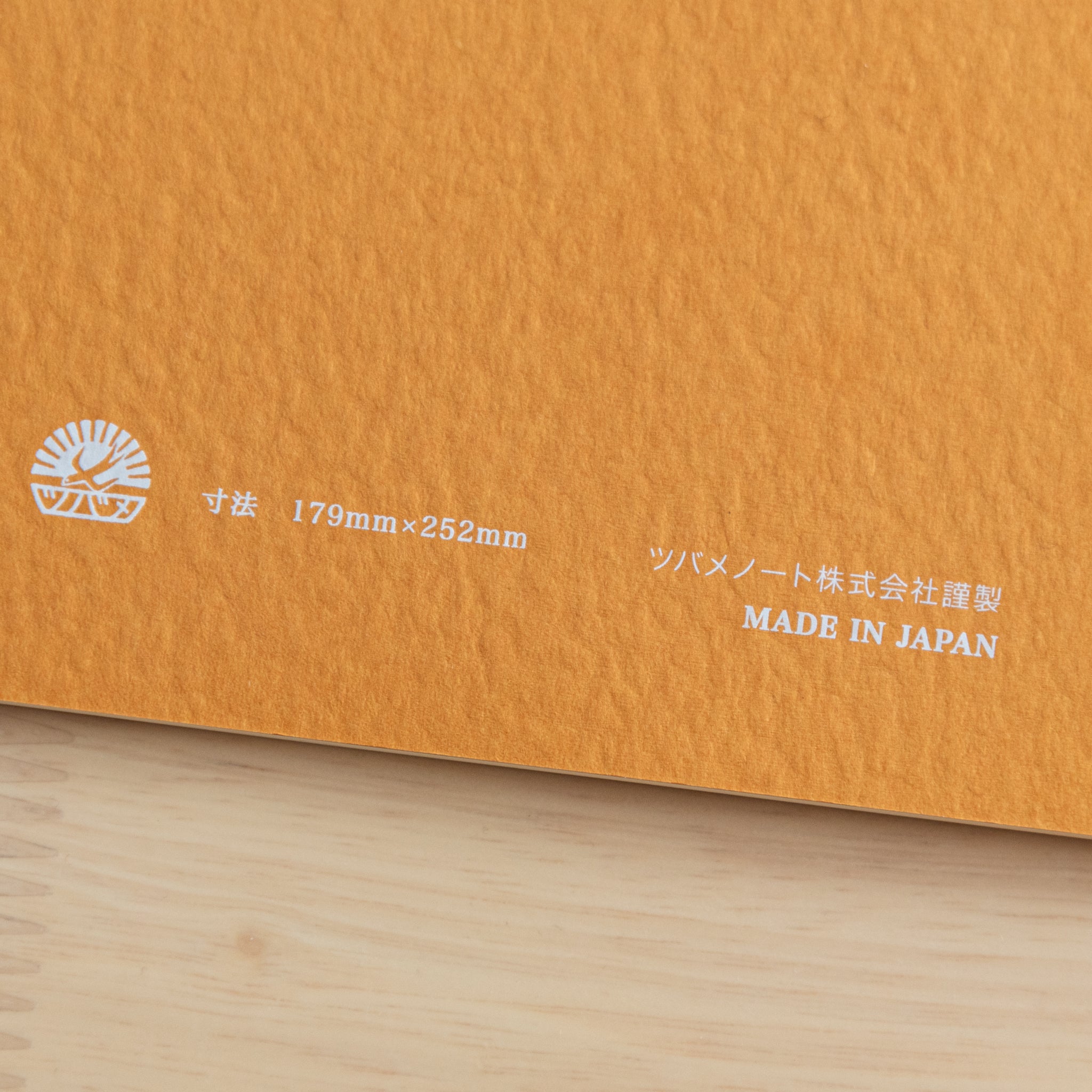 A close-up of the back cover of a Tsubame Japanese notebook, showing the logo, size details, and "Made in Japan" label on an orange textured surface.