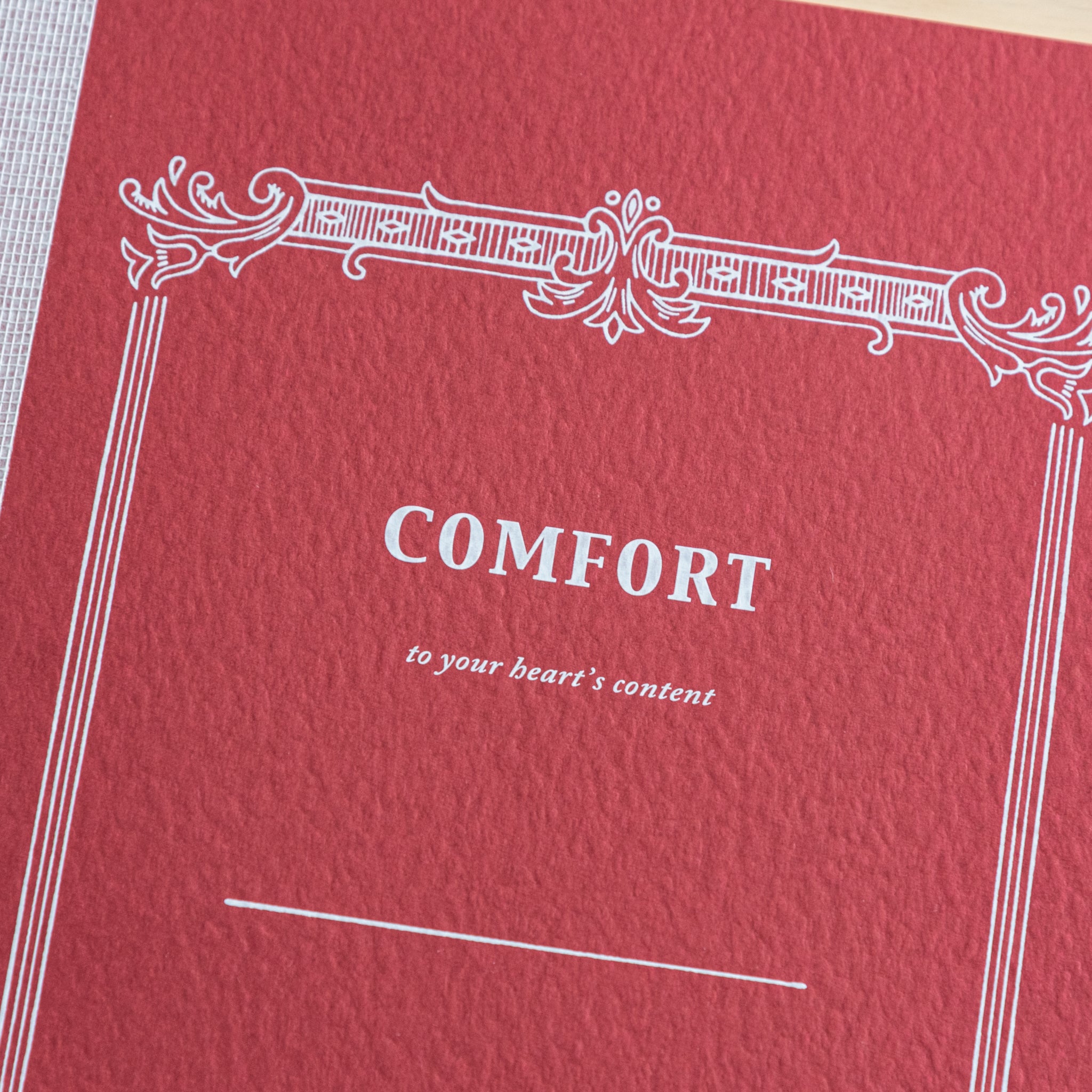 A close-up of the red cover of a Tsubame Japanese notebook labeled "Comfort," featuring an ornate vintage-style design. A refined piece of Japanese stationery.