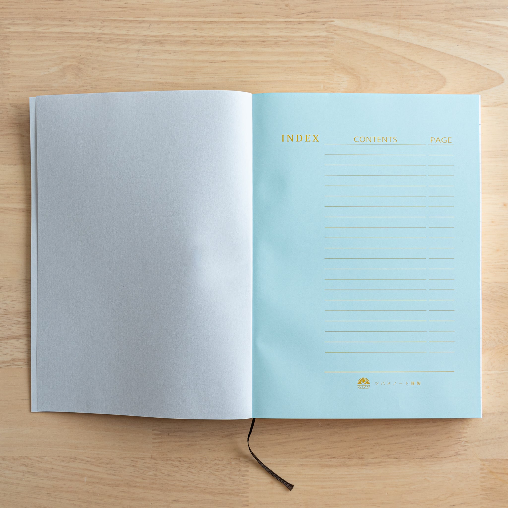 An open Japanese notebook displaying a light blue index page with gold lettering and a black ribbon bookmark, showcasing fine Japanese stationery.  