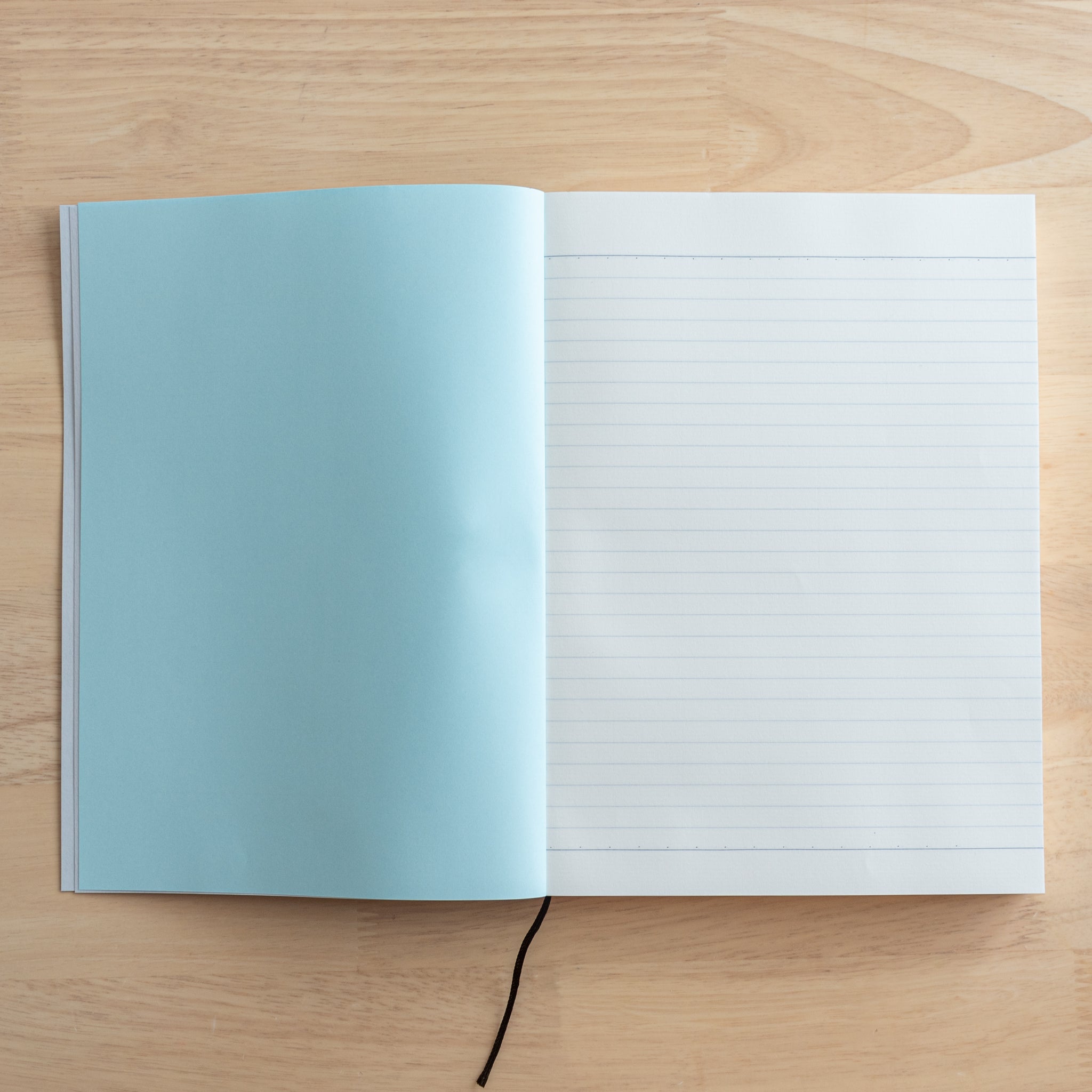 An open Japanese notebook showing a lined page with a light blue inner cover and a black ribbon bookmark, highlighting quality Japanese stationery.  