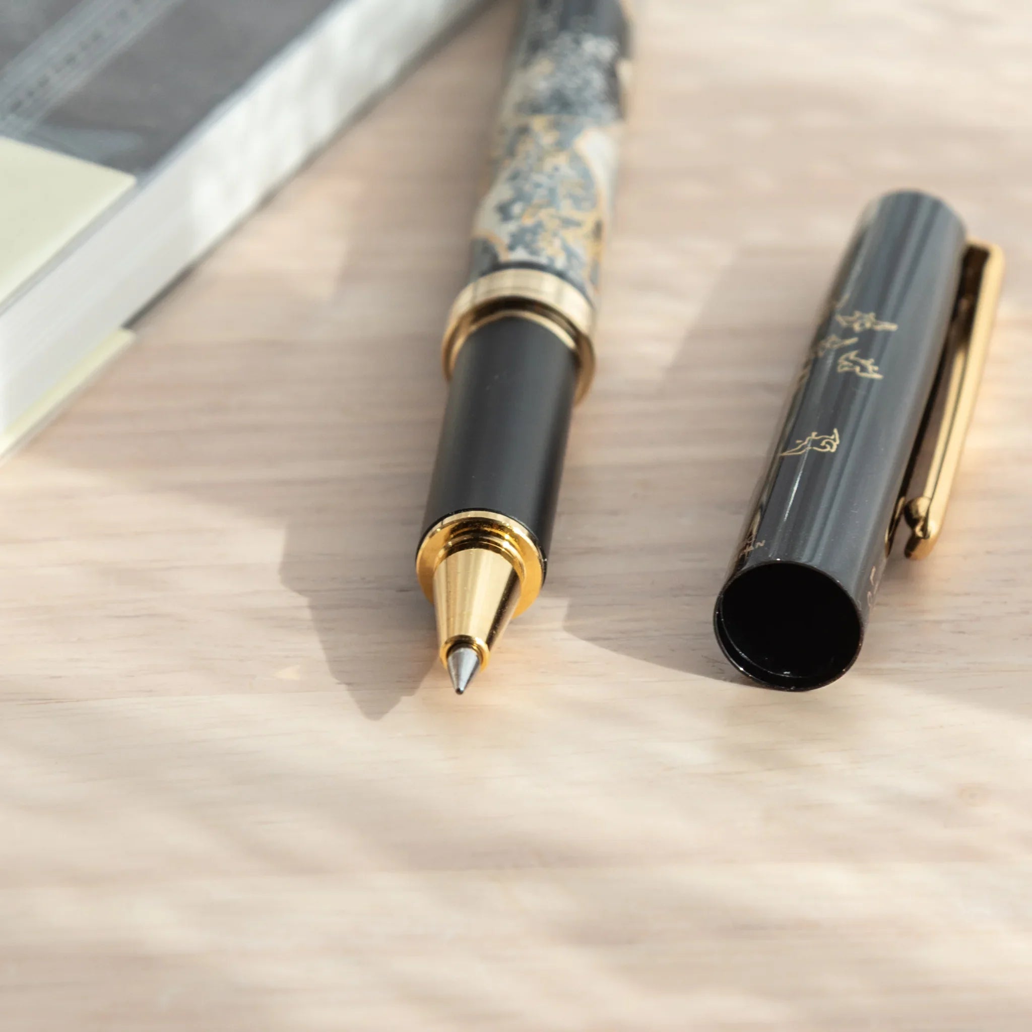 Maeju Yamanaka ballpoint pen body with Great Wave silkscreen and gold tip, beside black cap with gold clip, on wood surface