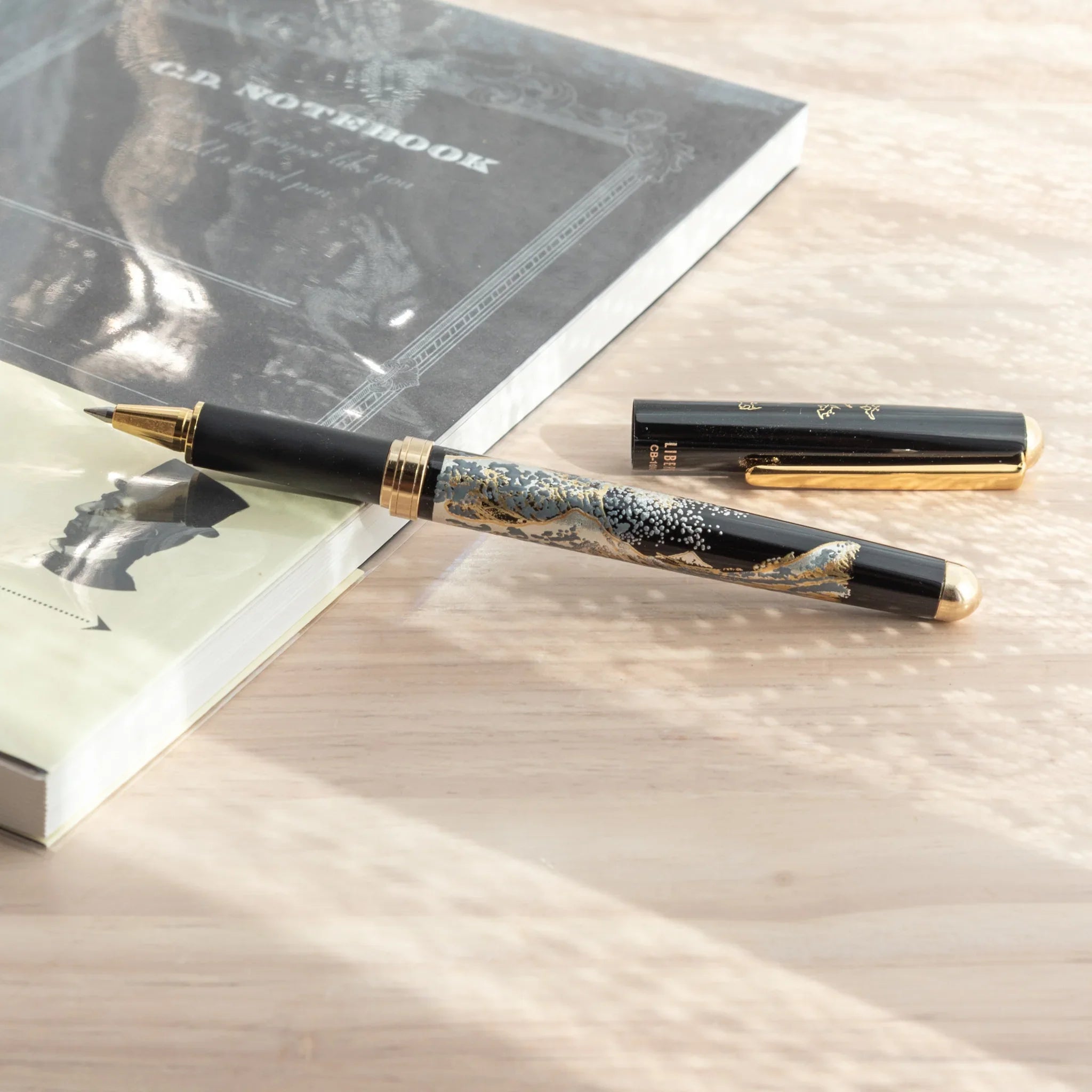 Maeju Yamanaka ballpoint pen body with Great Wave silkscreen and gold tip, cap separated beside dark notebook on wood surface