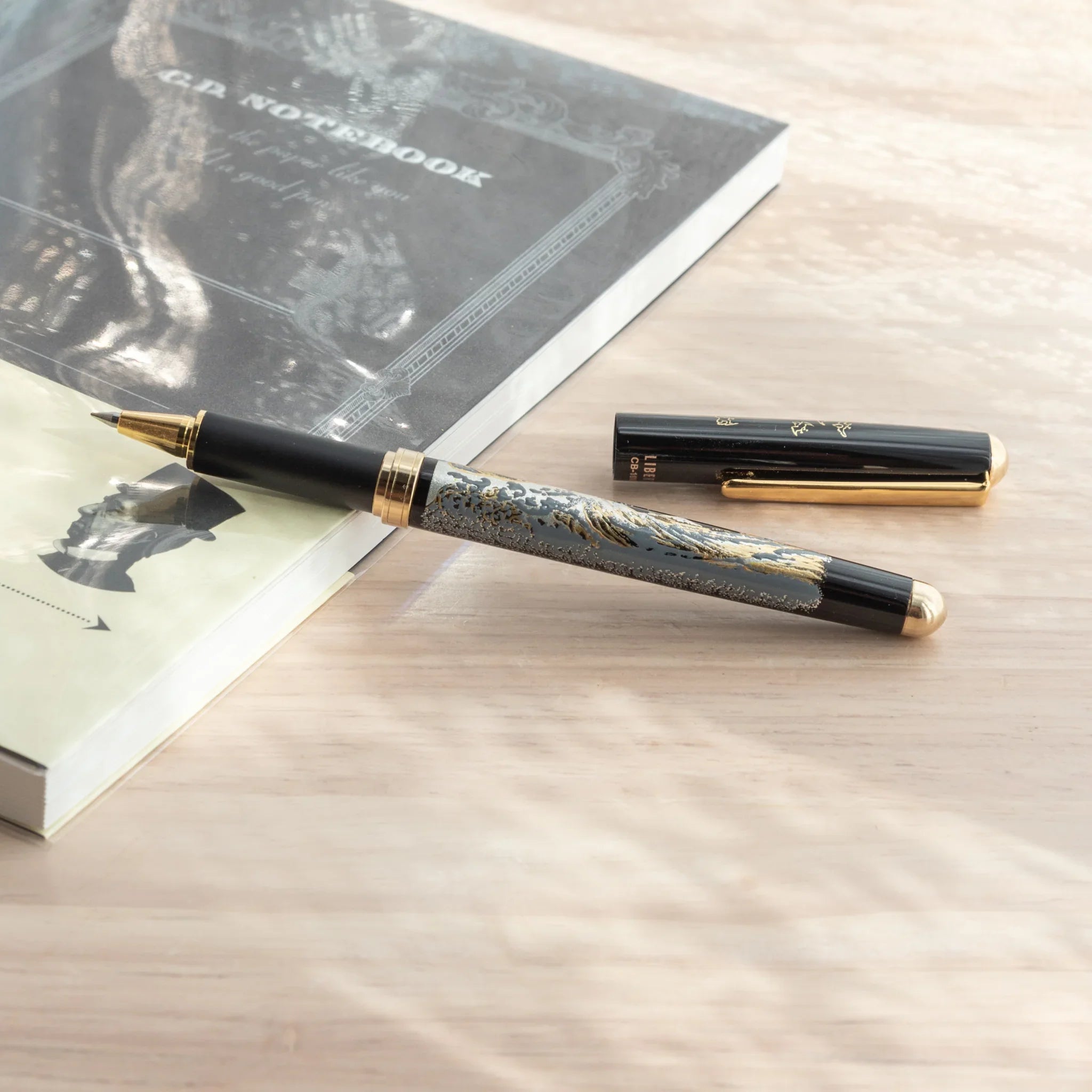 Maeju Yamanaka ballpoint pen and cap on wood surface showing Great Wave silkscreen detail, dark G.D. Notebook in background