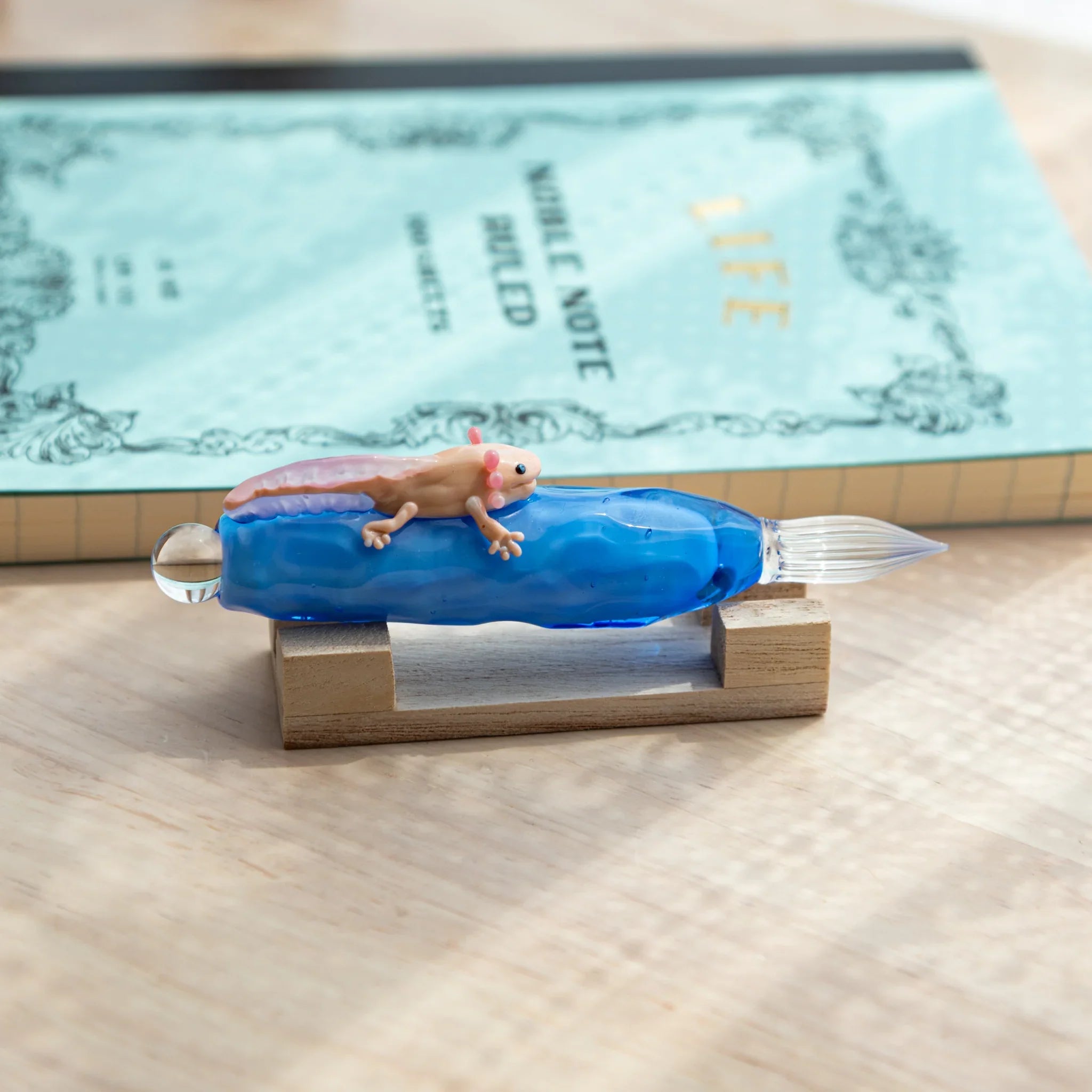 Blue glass dip pen with a tiny pink-gilled axolotl on the shaft, resting on a wooden stand beside a light blue notebook.