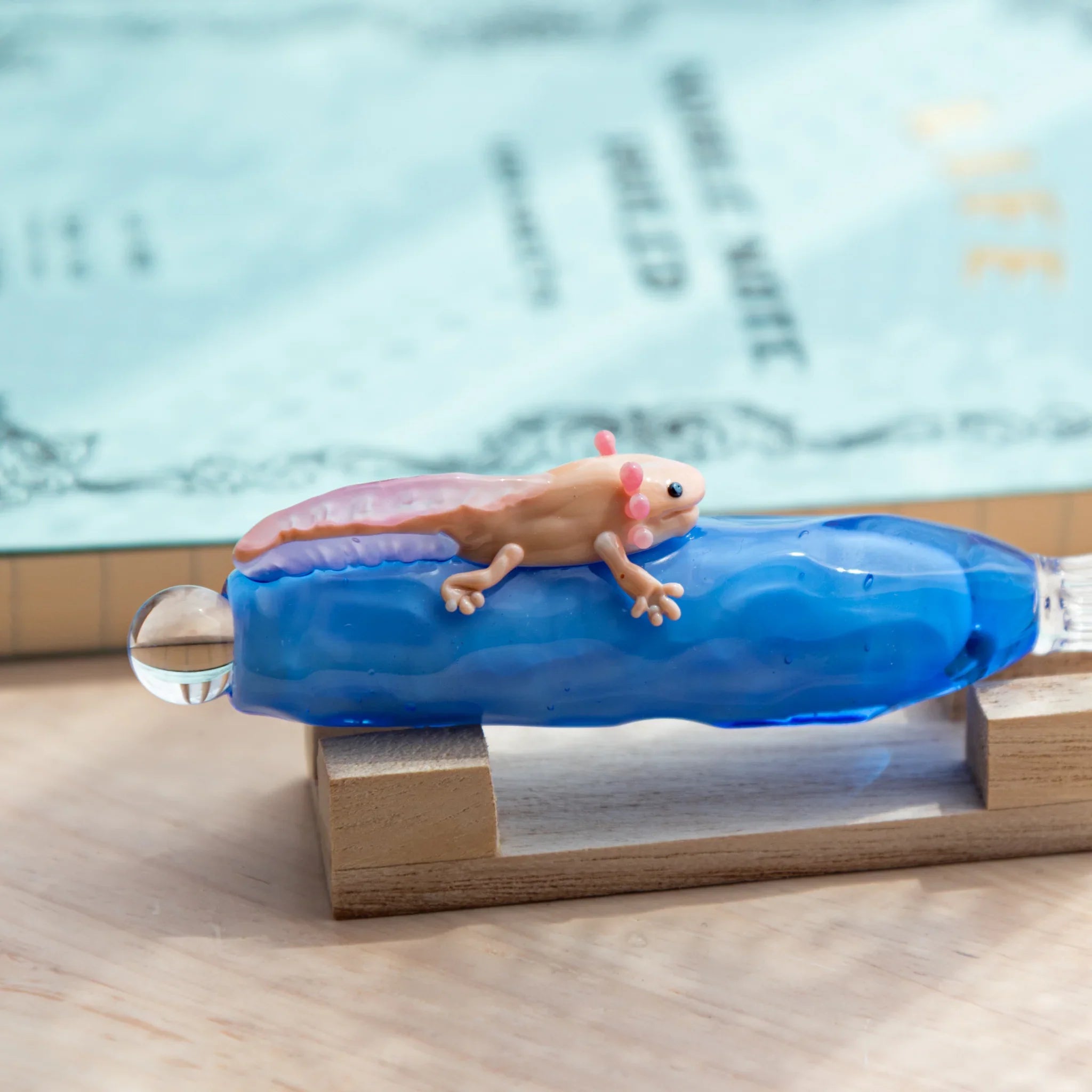 Blue glass dip pen with a tiny pink-gilled axolotl on the shaft, resting on a wooden stand beside a light blue notebook.