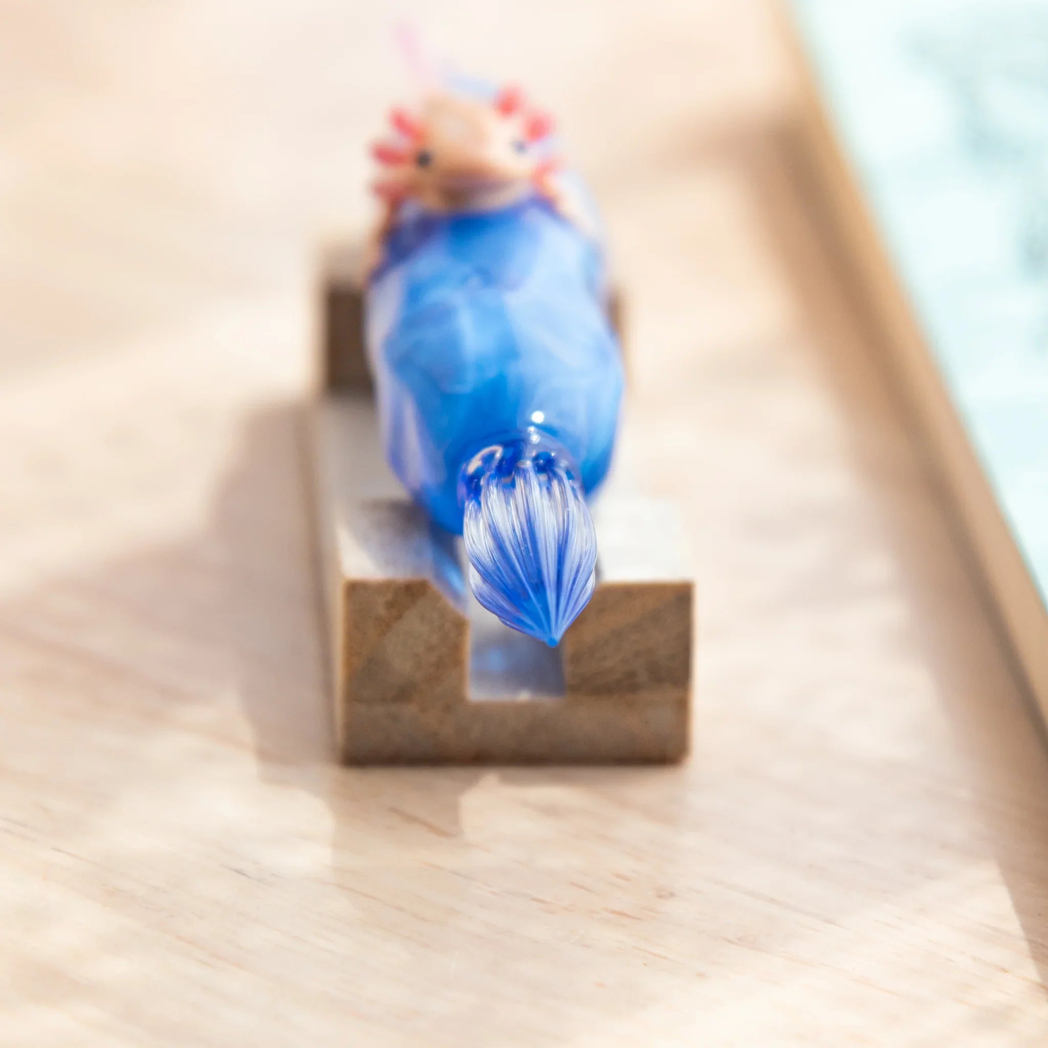 Close-up of a blue glass dip pen tip on a wooden stand, with a tiny pink-gilled axolotl figurine perched atop the shaft.