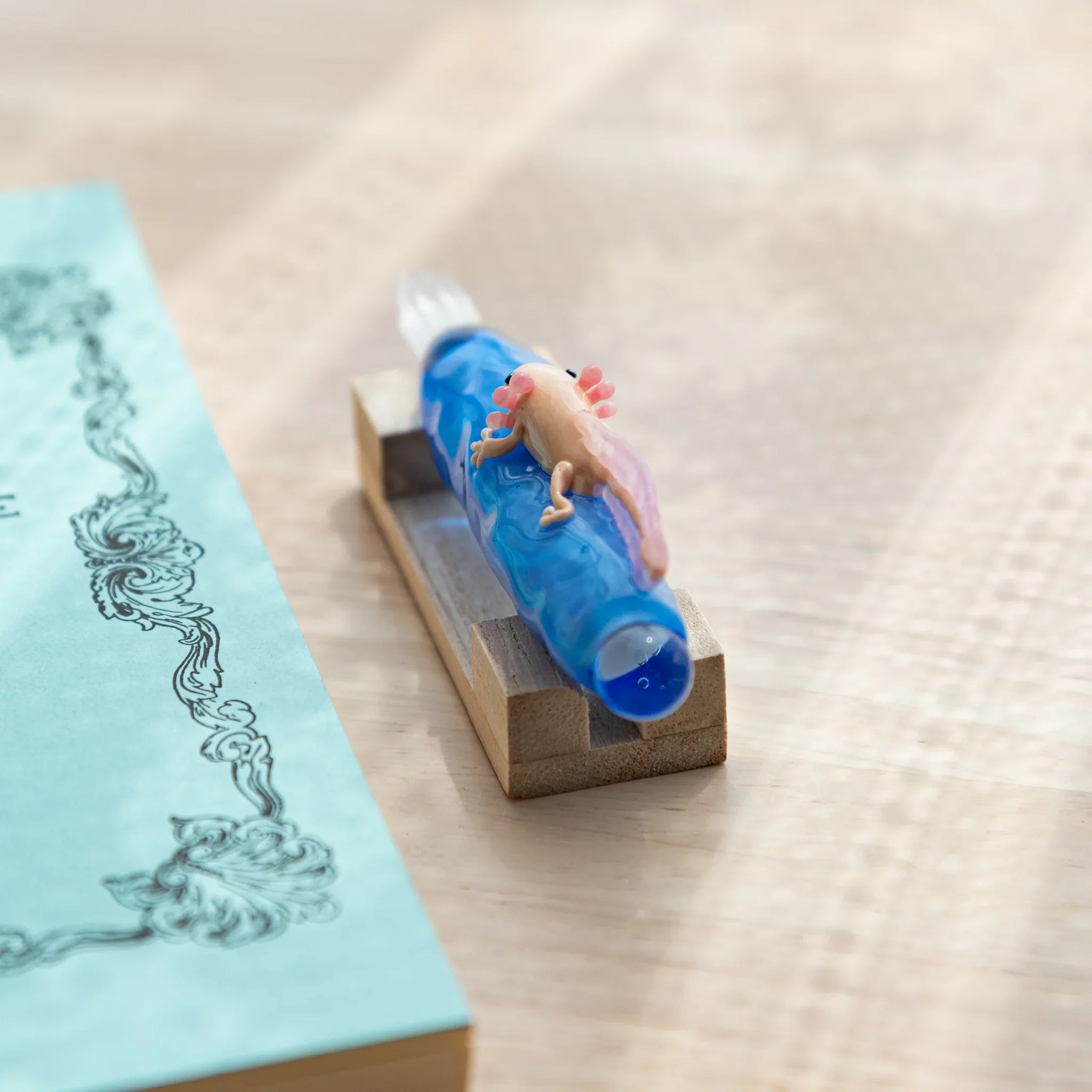 Blue glass dip pen with a small pink-gilled axolotl on the shaft, placed on a wooden stand beside a light blue ornate notebook.