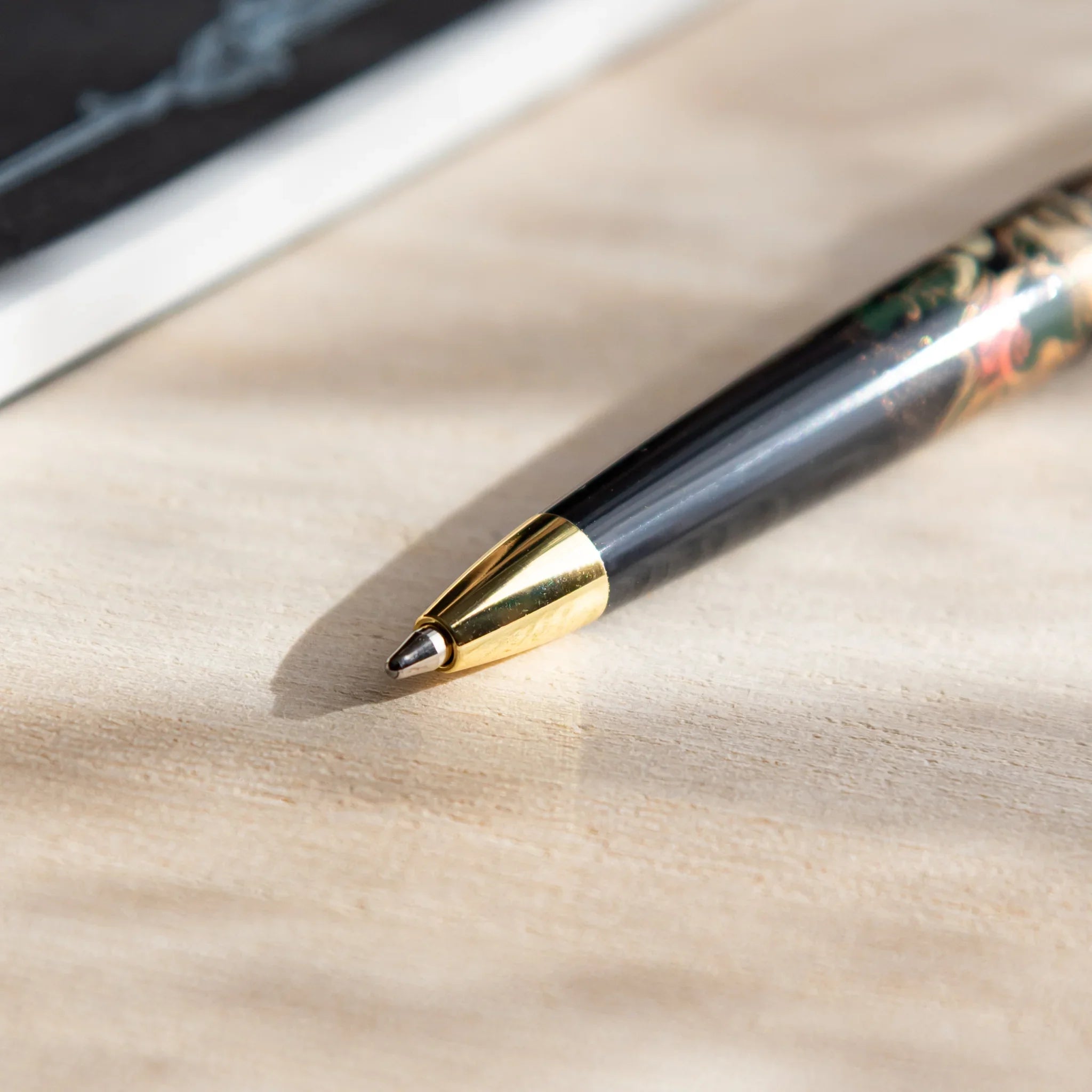 Close-up of Japanese Ballpoint Pen nib with gold tip resting on beige desk near a dark notebook.