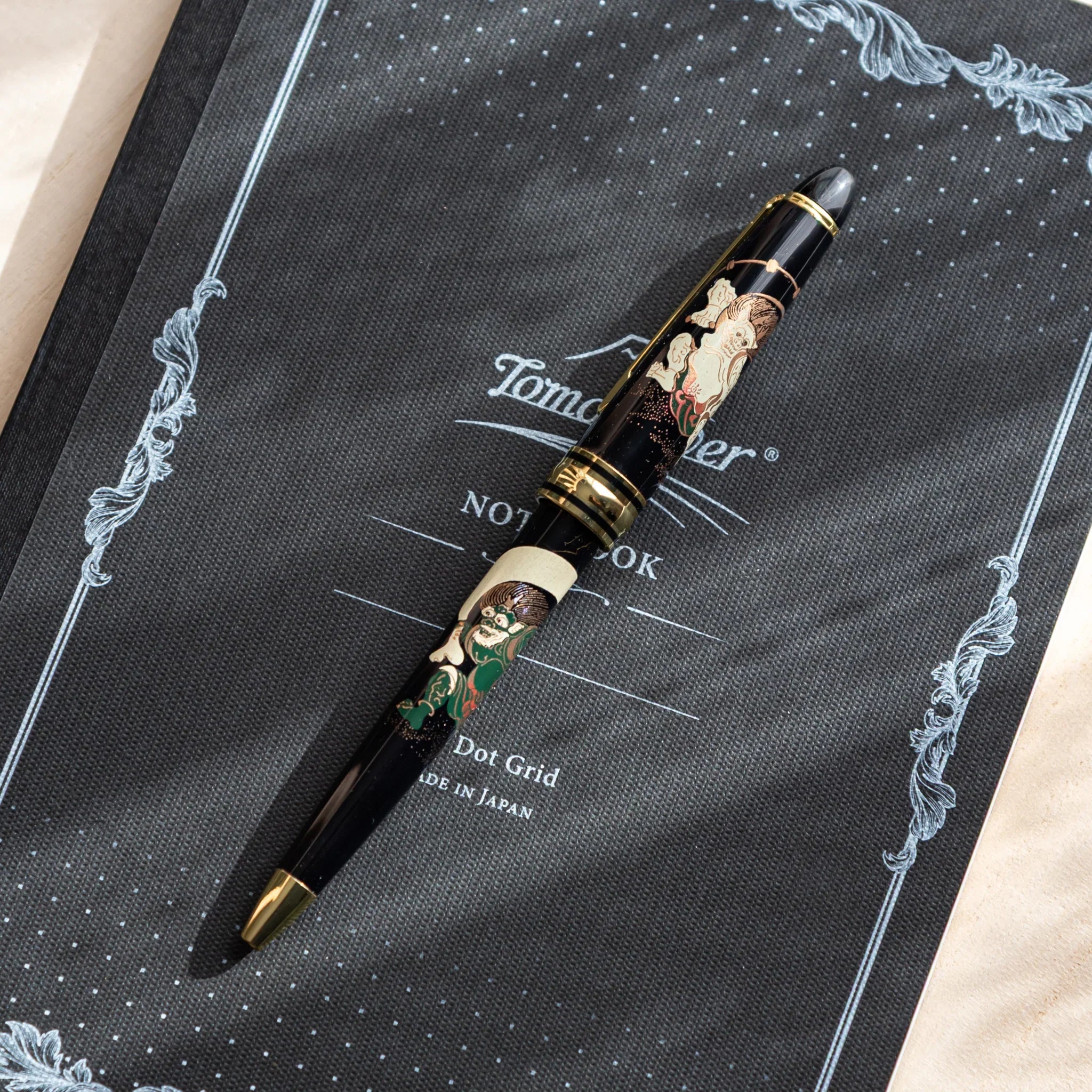 Japanese Ballpoint Pen with gold and black Raijin and Fujin design resting diagonally on dark patterned notebook.