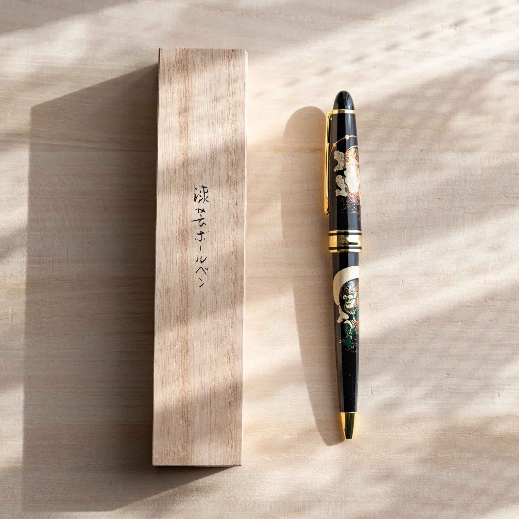 Japanese Ballpoint Pen with gold and black Raijin and Fujin design lying beside wooden box on light surface.