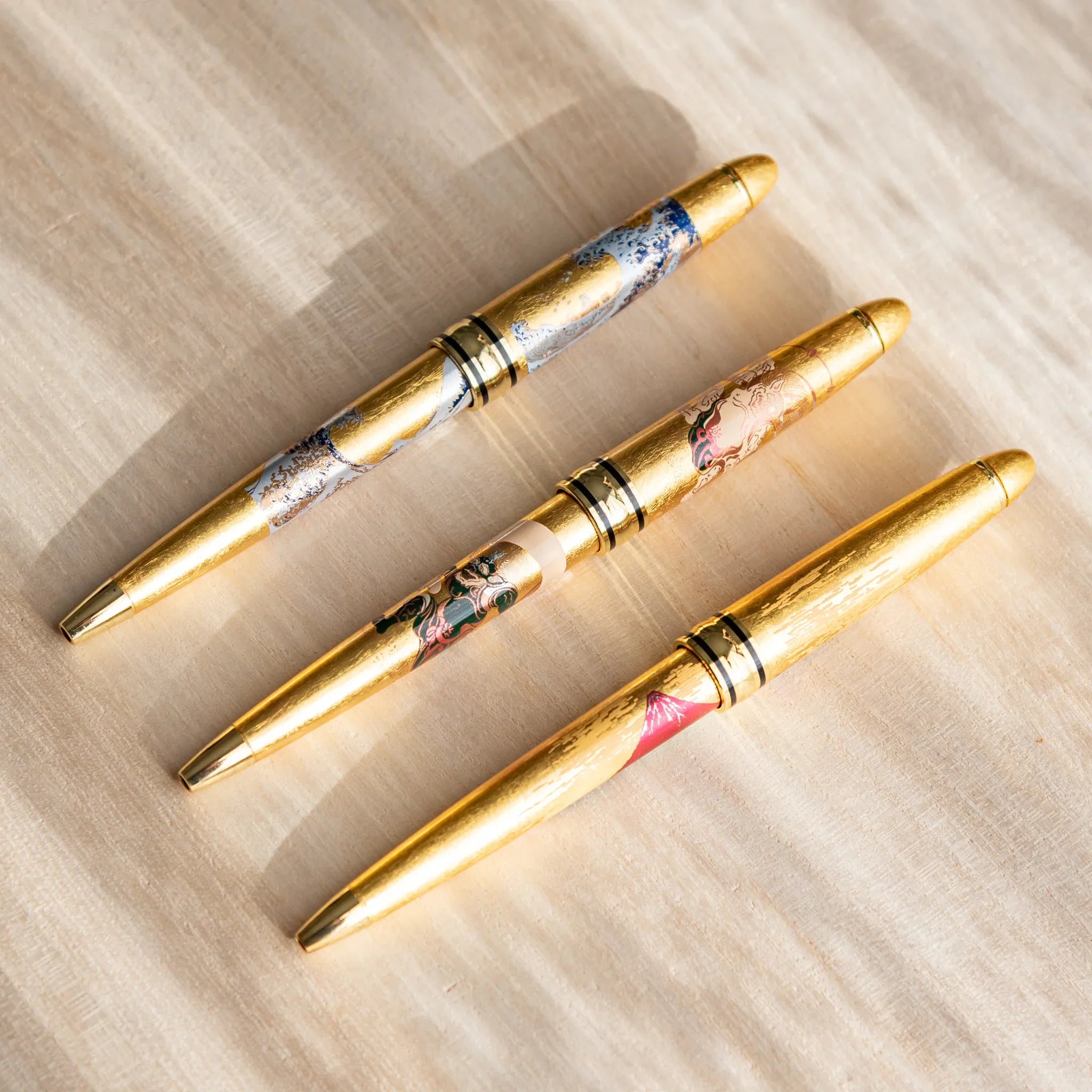 Three gold leaf Japanese Ballpoint Pens with Raijin and Fujin artwork displayed on a light wood surface.