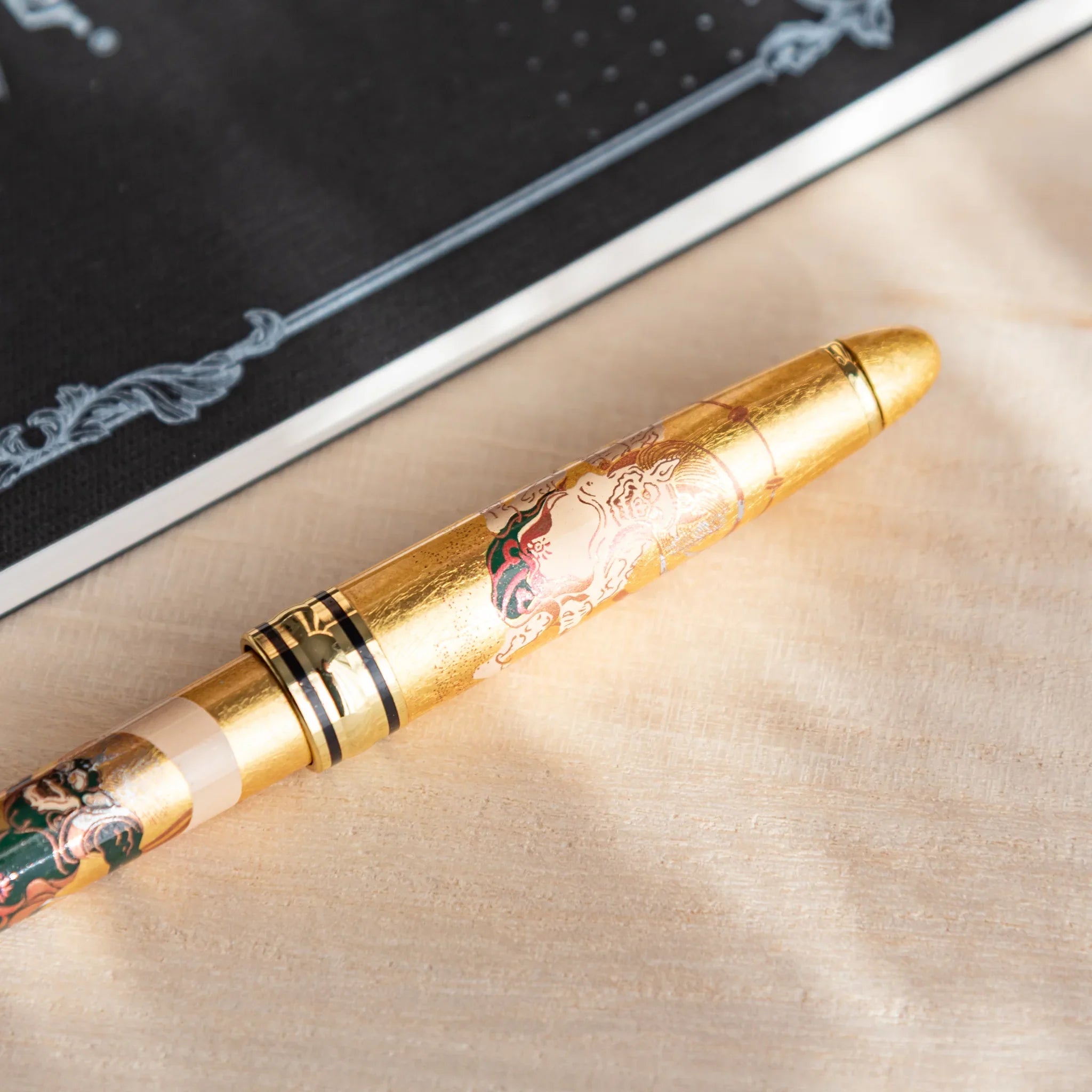 Gold leaf Japanese Ballpoint Pen featuring a detailed Fujin design, placed on a light wood desk beside a black notebook.