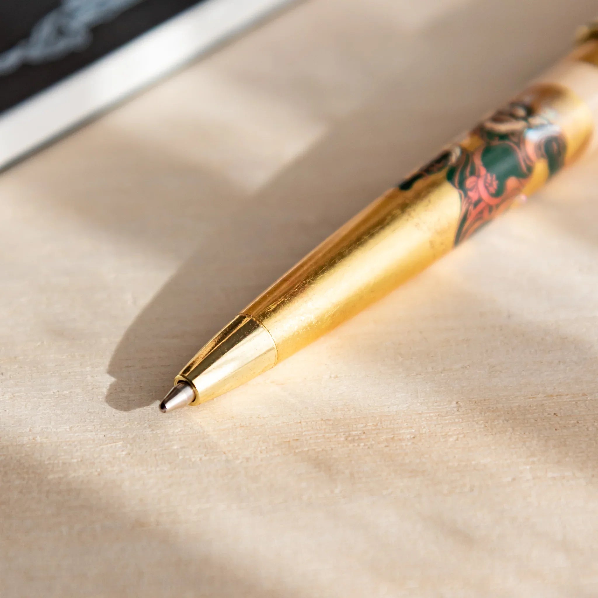 Close-up of a Japanese Ballpoint Pen with a gold tip and gold leaf Raijin and Fujin design, resting on a light wood desk.