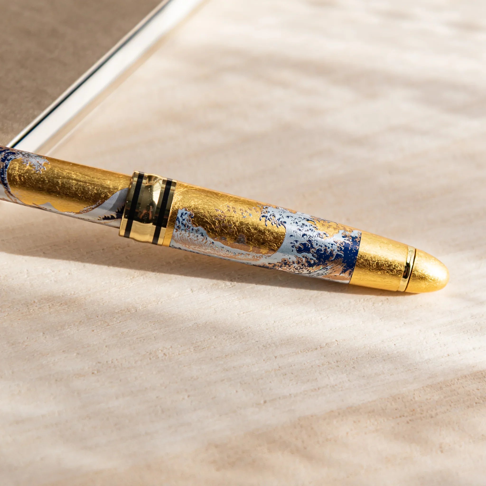 Gold Leaf Great Wave Japanese Ballpoint Pen on a light wood desk, showing blue-and-gold wave details and a shining gold cap.