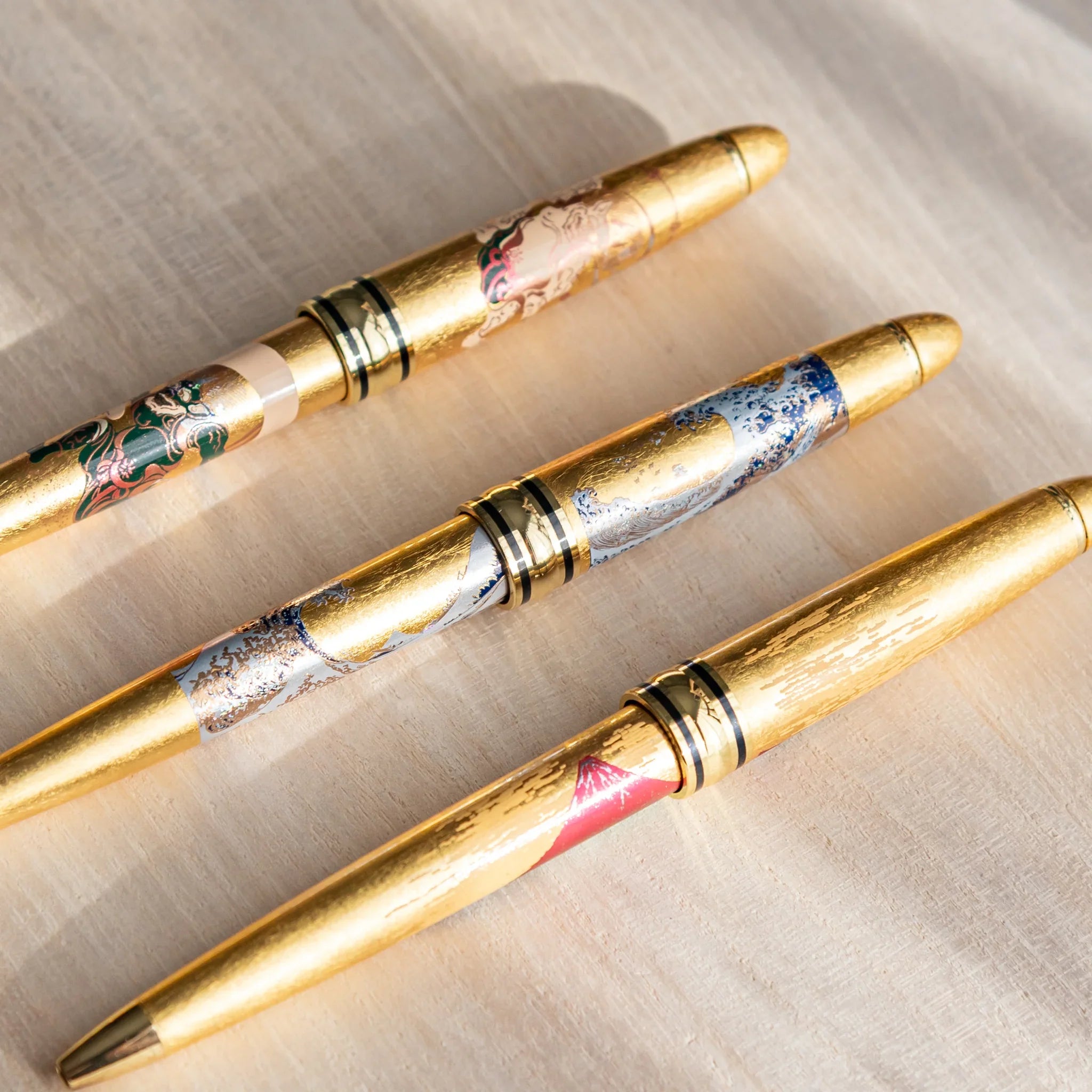 Three gold leaf Japanese Ballpoint Pens arranged diagonally on a desk, showing wave, Fujin, and Raijin designs with gold bodies.