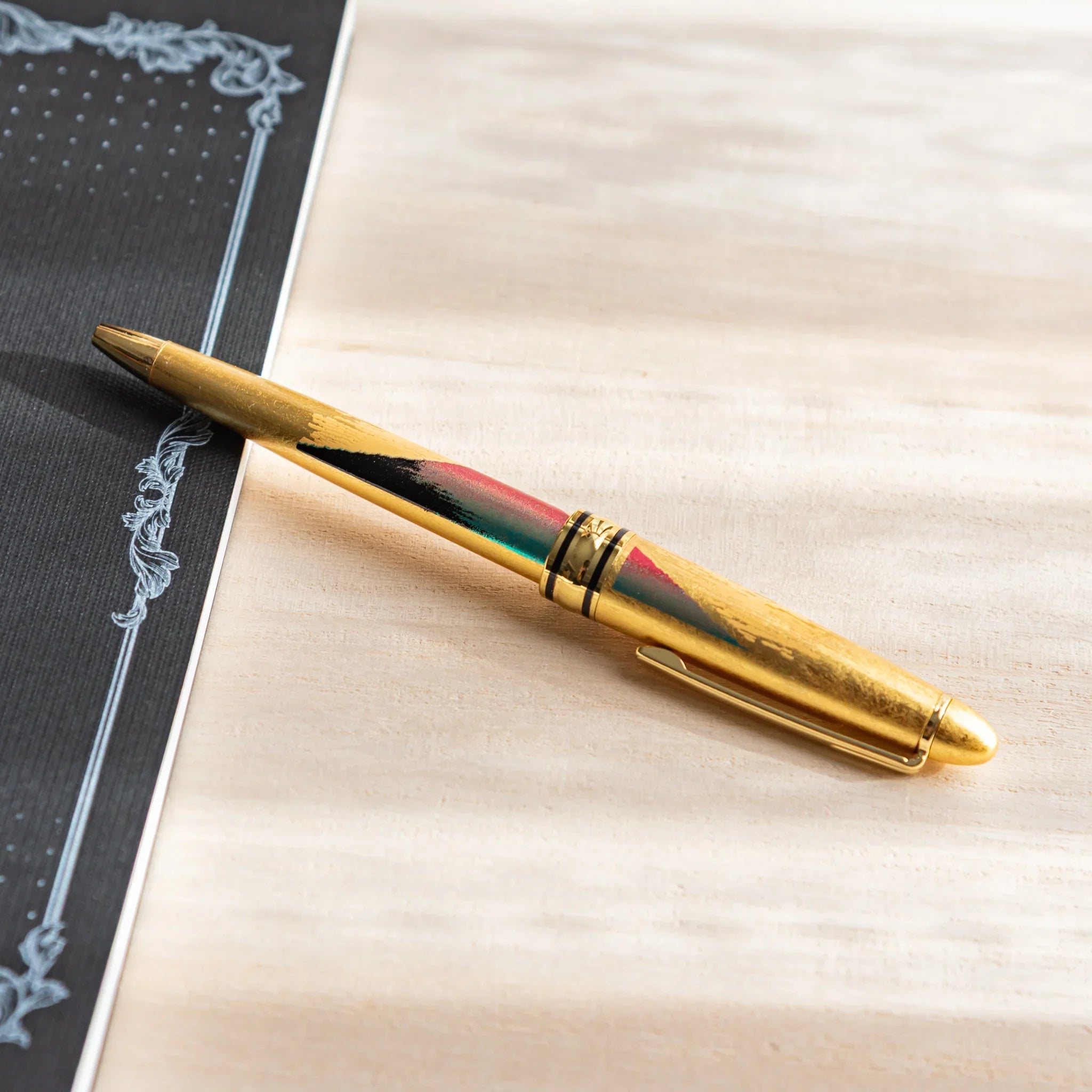 Gold Leaf Mt. Fuji Japanese Ballpoint Pen on a desk beside a black notebook, showing red Fuji detail and a gold body.