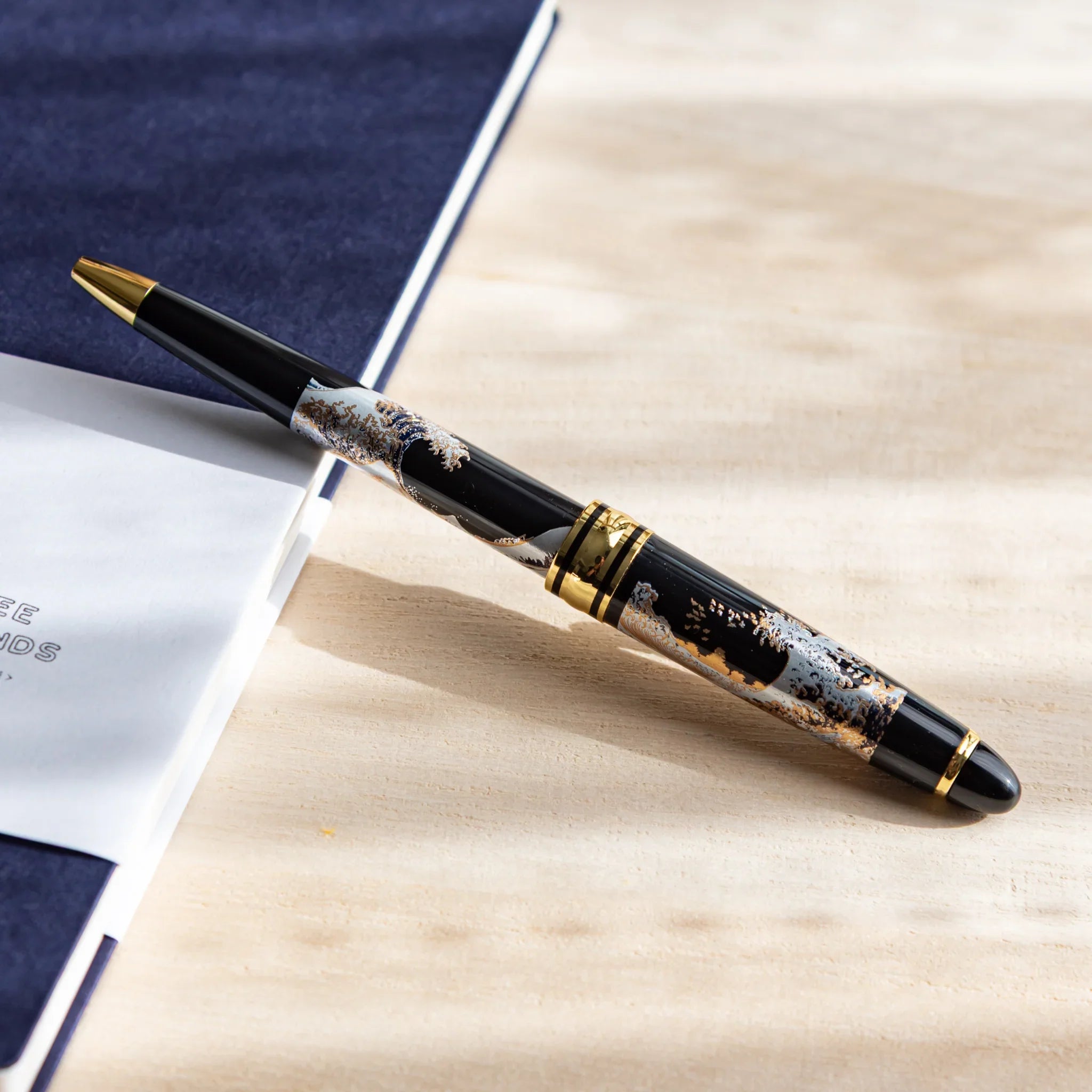 Japanese Ballpoint Pen with Great Wave design placed diagonally on navy notebook, showing black lacquer body.