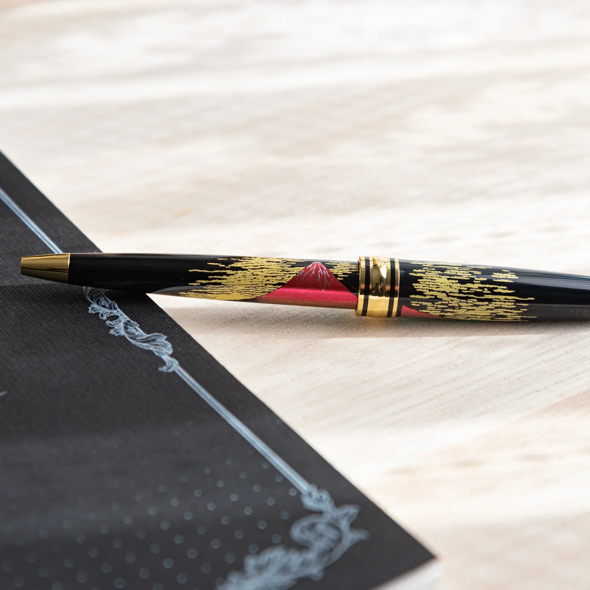 Japanese Ballpoint Pen with red Mt. Fuji and gold cloud design on a light wood desk beside a black notebook.