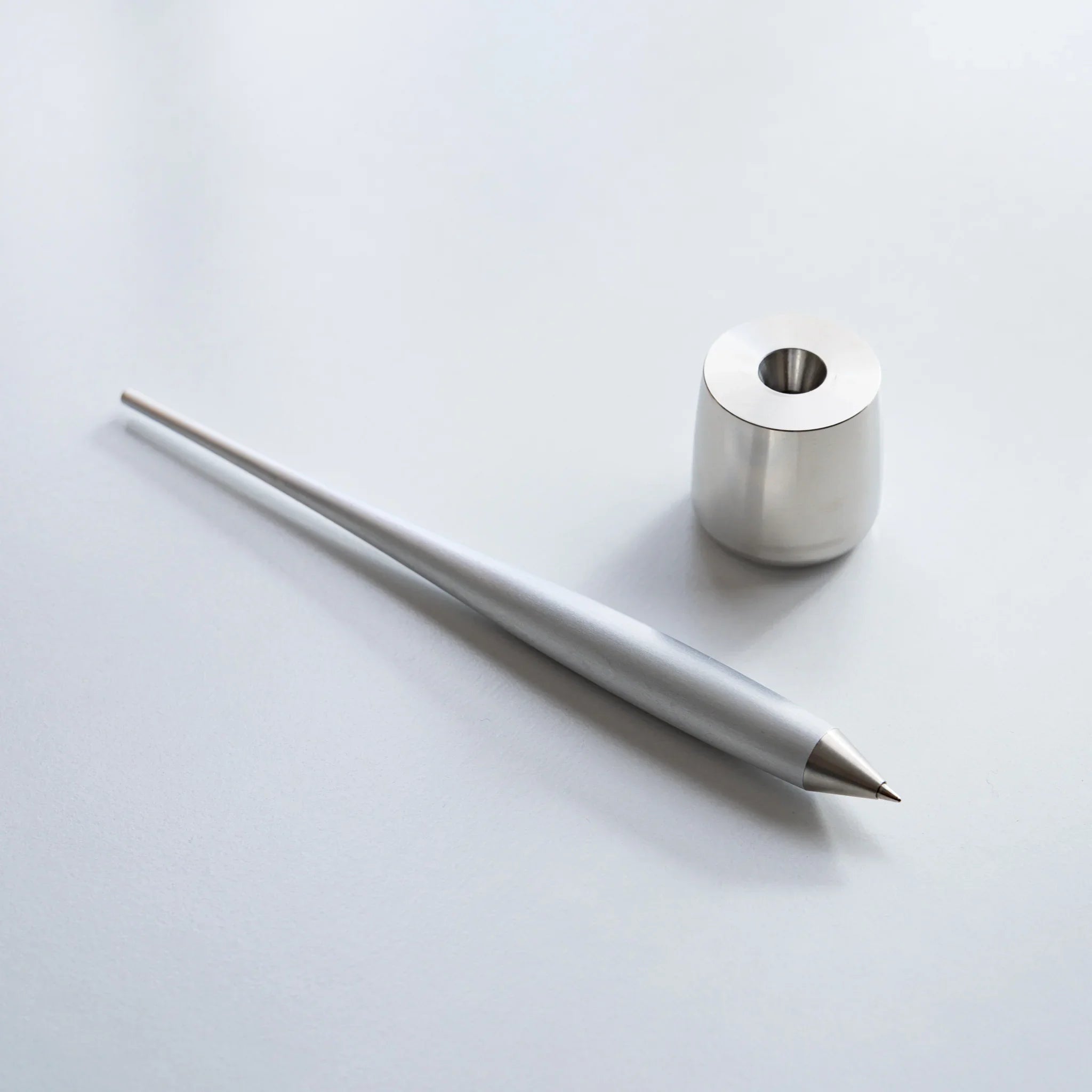 Duralumin ballpoint pen resting diagonally next to brushed stainless steel cylindrical pen stand holder base