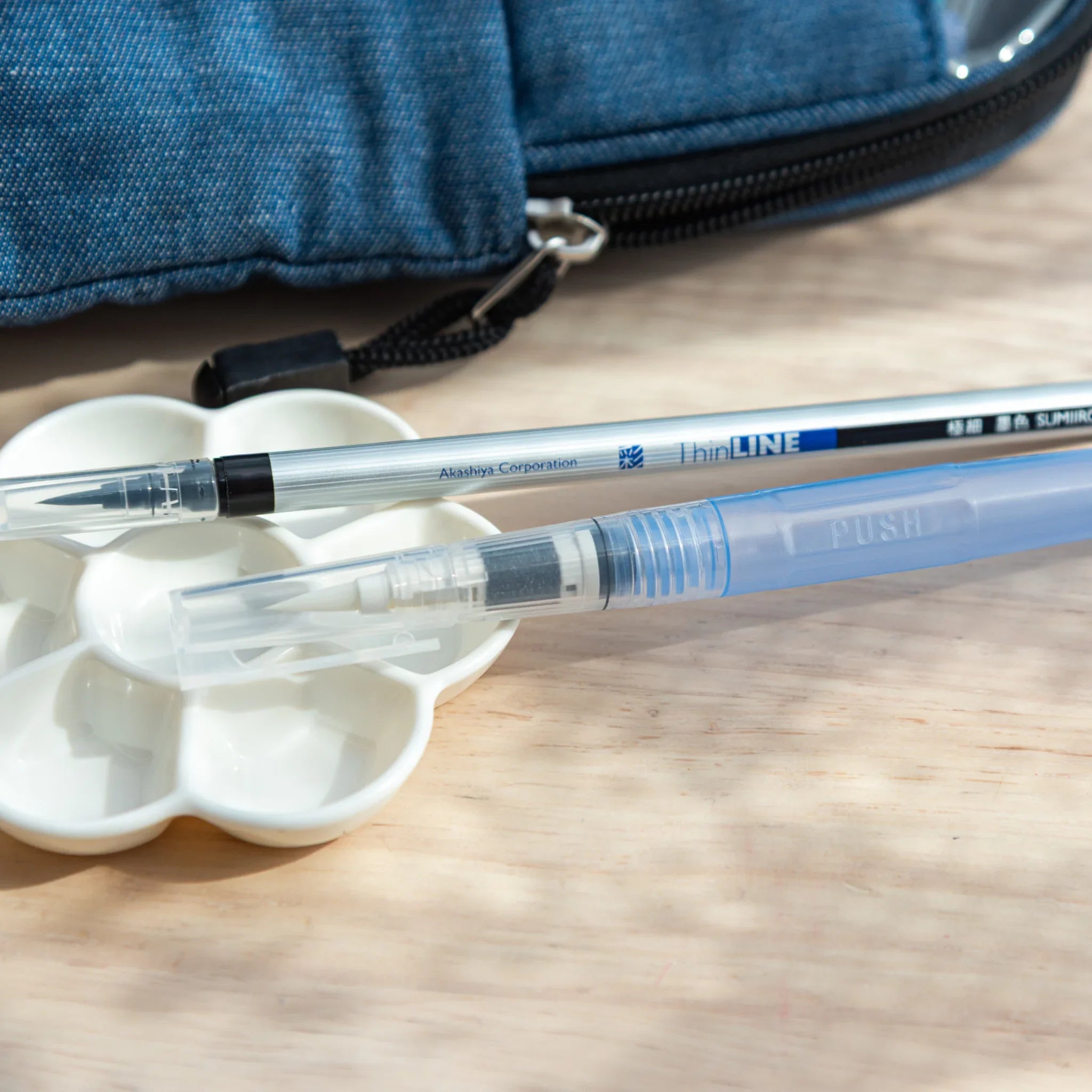 Water brush and sumi ink brush placed on a white palette with denim case in background, for arts and crafts sketching.