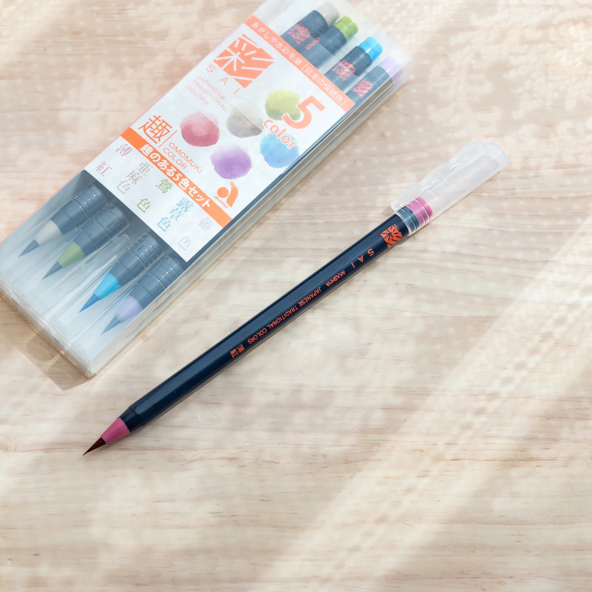 A single Akashiya Sai Japanese Brush Pen in pink lies beside the full set on a wooden desk.