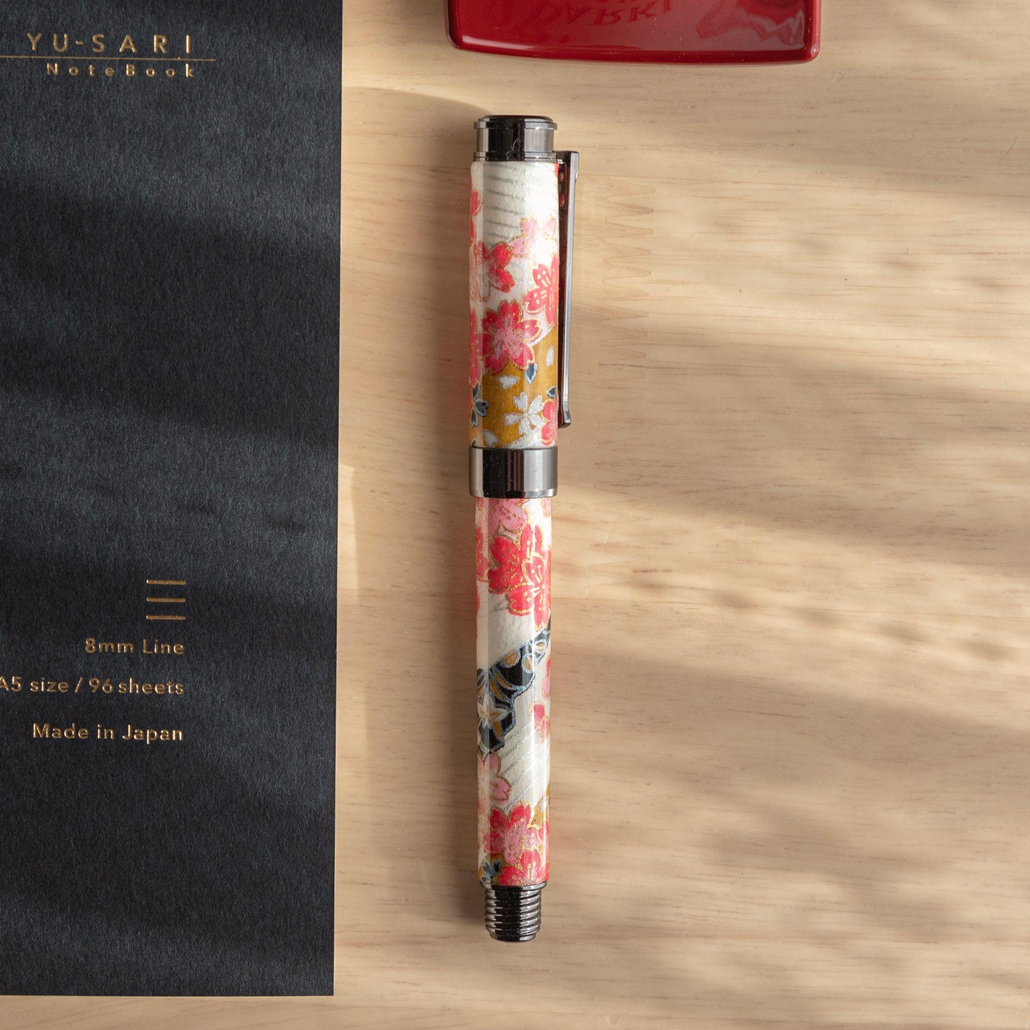 Floral-patterned fountain pen with cherry blossoms on a washi finish, placed beside a black notebook embossed with gold text, on a wooden surface.