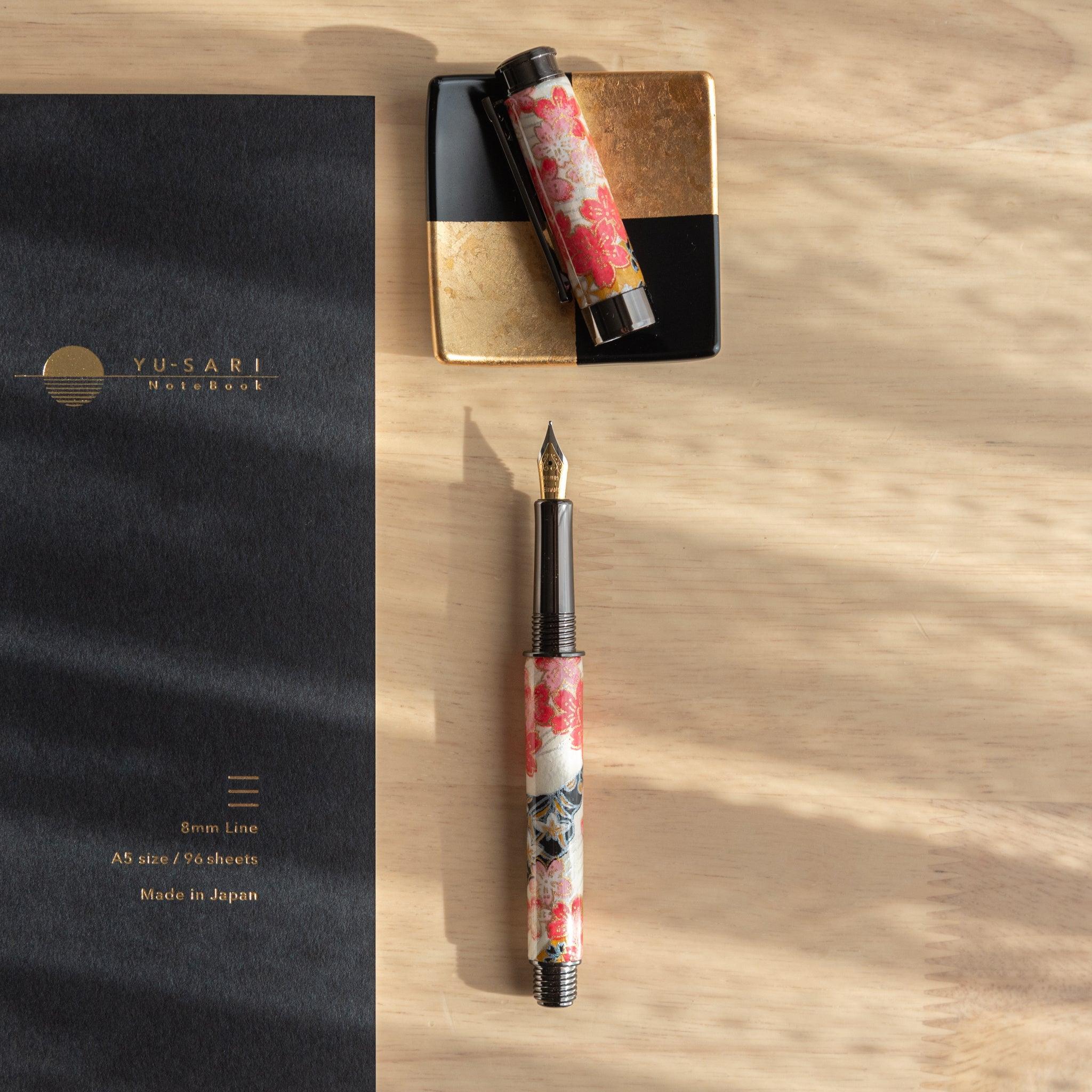 Fountain pen with cherry blossom design and gold nib placed beside its cap on a golden pen holder, next to a black "Yu-Sari" notebook.