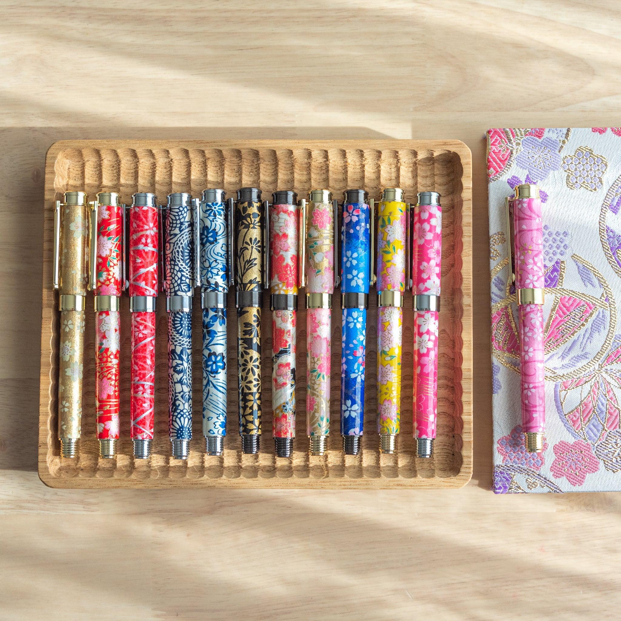 Collection of vibrant washi-patterned fountain pens arranged in a wooden tray, featuring floral and traditional motifs, on a light wooden desk.