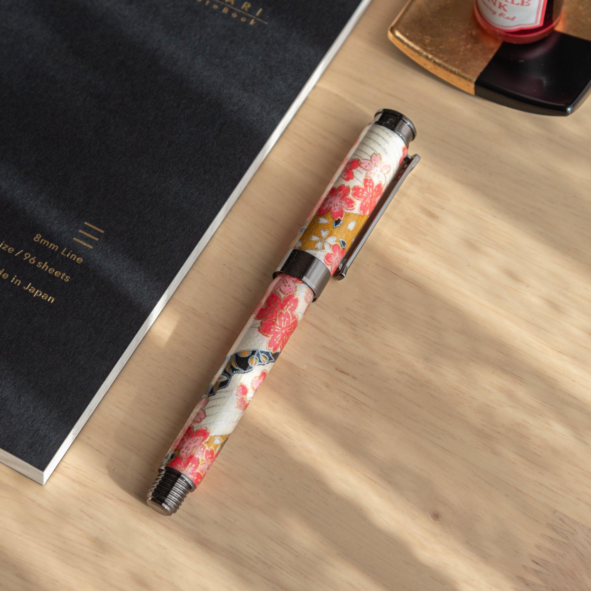 Floral fountain pen with cherry blossom motifs, resting diagonally on a black "Yu-Sari" notebook, highlighted by soft natural light.