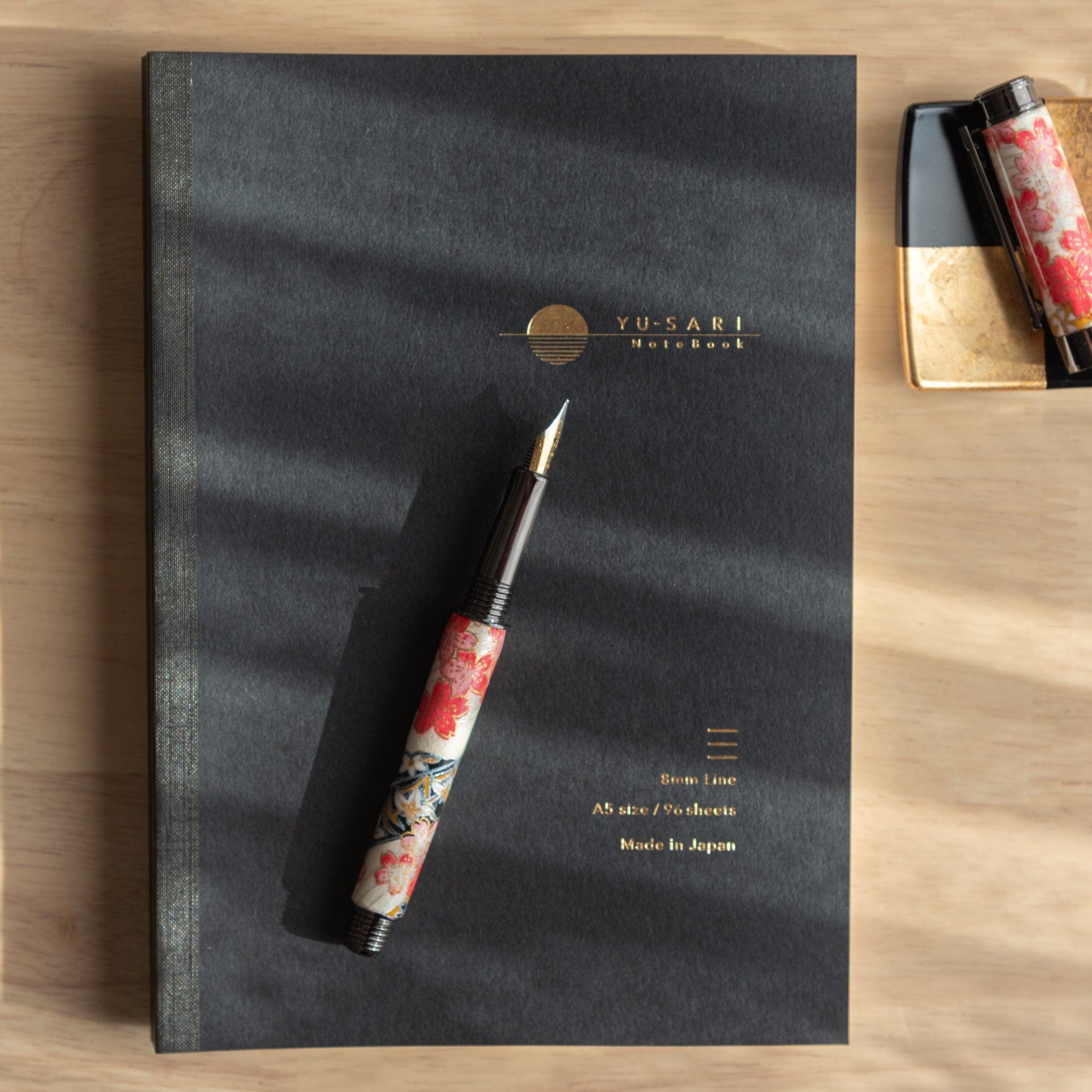 Opened floral fountain pen on a black "Yu-Sari" notebook, with golden text visible, alongside an ink bottle on a wooden desk.